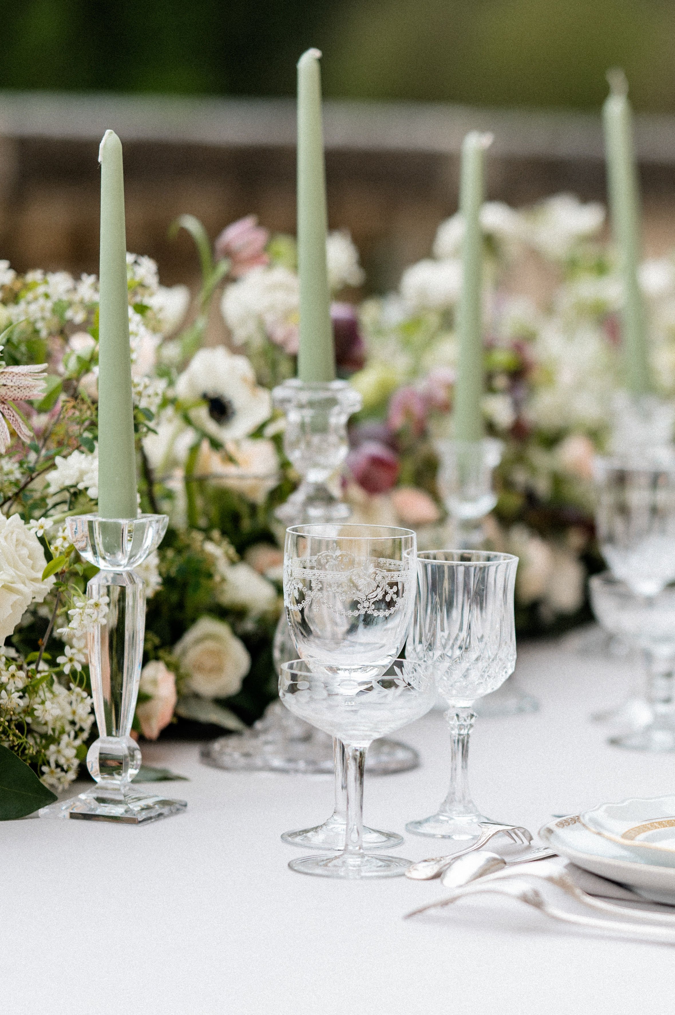 A close-up detail shot of an outdoor wedding reception table set with a white linen. The centerpiece features a lush low floral arrangement mixing white roses, white anemones, blush ranunculus, mauve blooms, and greenery, interspersed with sage green taper candles in clear crystal candlestick holders. In the foreground, two etched crystal wine glasses and a cut-crystal wine glass are arranged at a place setting alongside silver flatware and white plates with a gold rim accent. The overall decor palette is white, blush, mauve, and sage green, with a classic French-inspired styling.