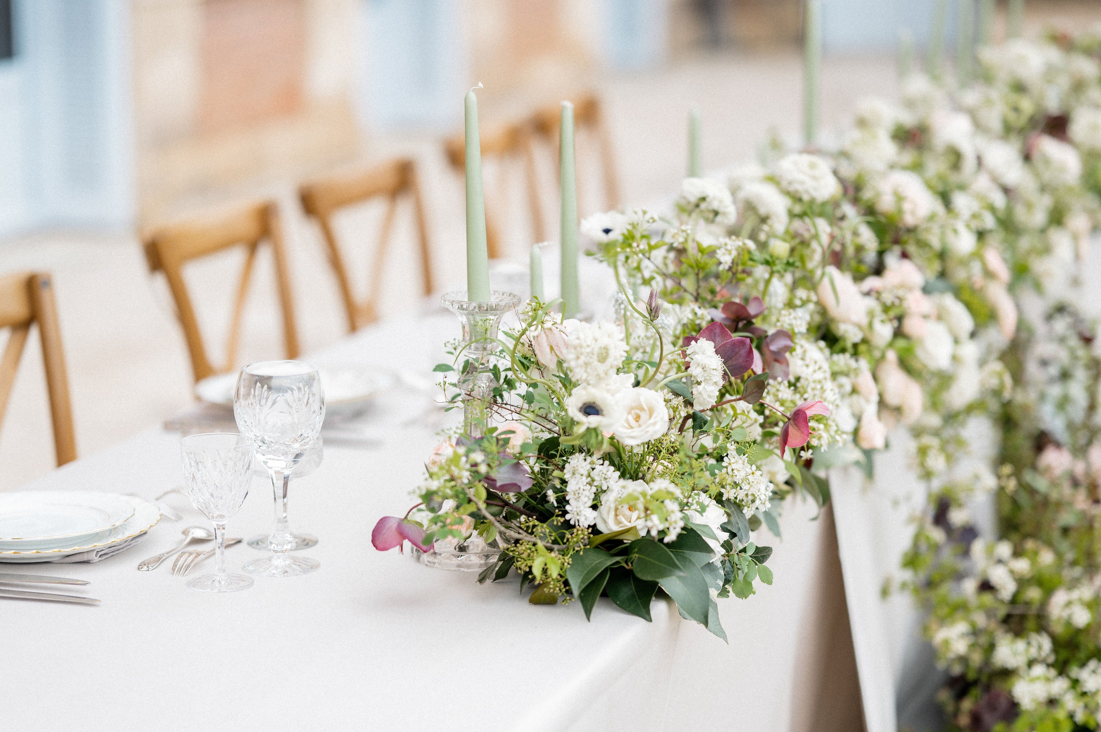 A close-up detail shot of a wedding reception tablescape, showing a long white linen-covered table set for a meal. The centerpiece features a lush floral runner composed of white roses, white anemones with dark centers, white clustered blooms resembling lilac or spirea, blush tulips, burgundy foliage, and abundant green trailing foliage. Sage green taper candles in crystal candleholders are integrated into the floral arrangement at varying heights. The place settings include stacked white china plates with a gold rim detail, crystal cut-glass wine glasses, and silver cutlery. Natural wood cross-back chairs are visible in the soft-focus background, and the overall decor palette combines white, sage green, blush, and burgundy in a garden-inspired classic style.