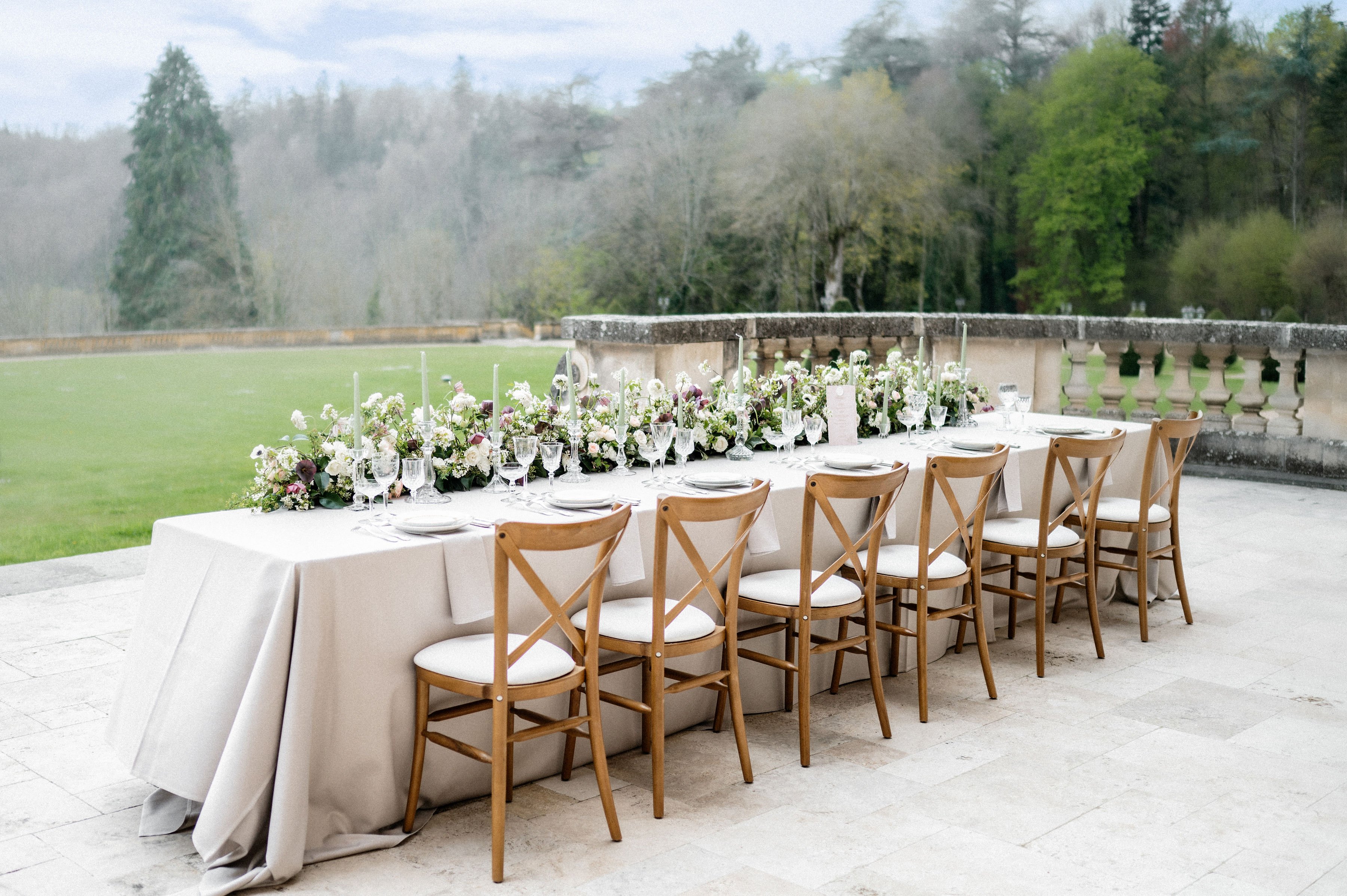 An outdoor reception tablescape set on a stone terrace alongside a carved balustrade railing, with no people present. The long rectangular table is dressed in a light greige linen tablecloth and lined with natural wood cross-back chairs with cream upholstered seat pads. A lush floral garland runner stretches the full length of the table, featuring ivory garden roses, white anemones, deep plum anemones, mauve blooms, and trailing greenery. Tall taper candles in soft grey tones are interspersed throughout the runner, and crystal wine glasses, grey charger plates, and white napkins complete each place setting. The overall decor palette is greige, ivory, and dusty plum with a classic, romantic styling. Wide shot taken from a slightly elevated angle along the side of the table.