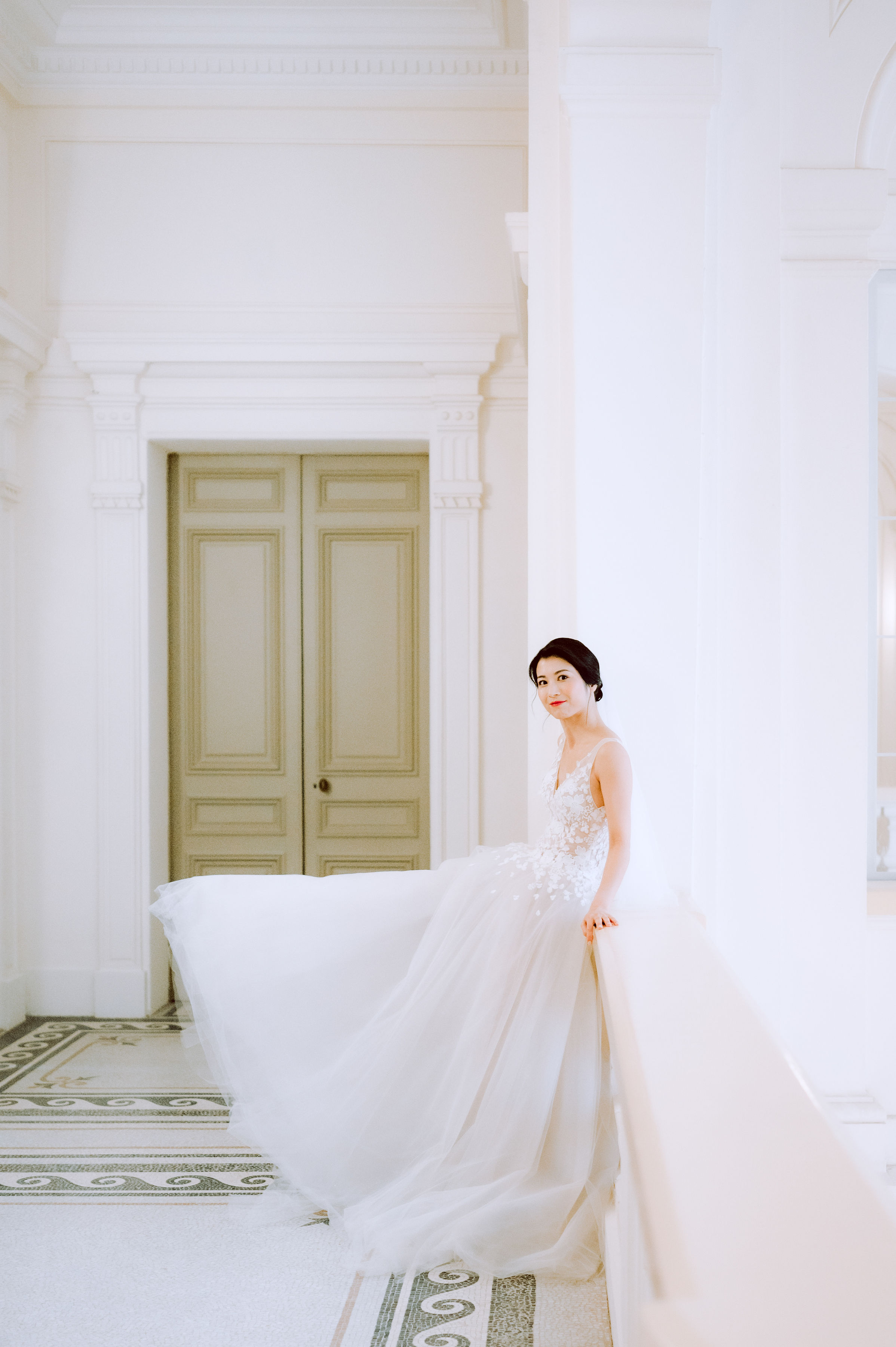A bridal portrait taken indoors in a grand classical interior featuring white molded walls, ornate cornicing, and an intricate mosaic tile floor in green and white patterns. The bride stands beside a large white pillar, leaning slightly against it and looking back toward the camera, with her voluminous ivory tulle skirt billowing outward to fill much of the frame. She wears a fitted lace-bodice gown with thin straps and a deep V-neckline, with her dark hair styled in a low updo and a red lip as the sole accent color. A pair of olive-green painted double doors with carved paneling frames the background. The composition is a three-quarter portrait shot with bright, airy natural light creating a high-key, low-contrast mood.