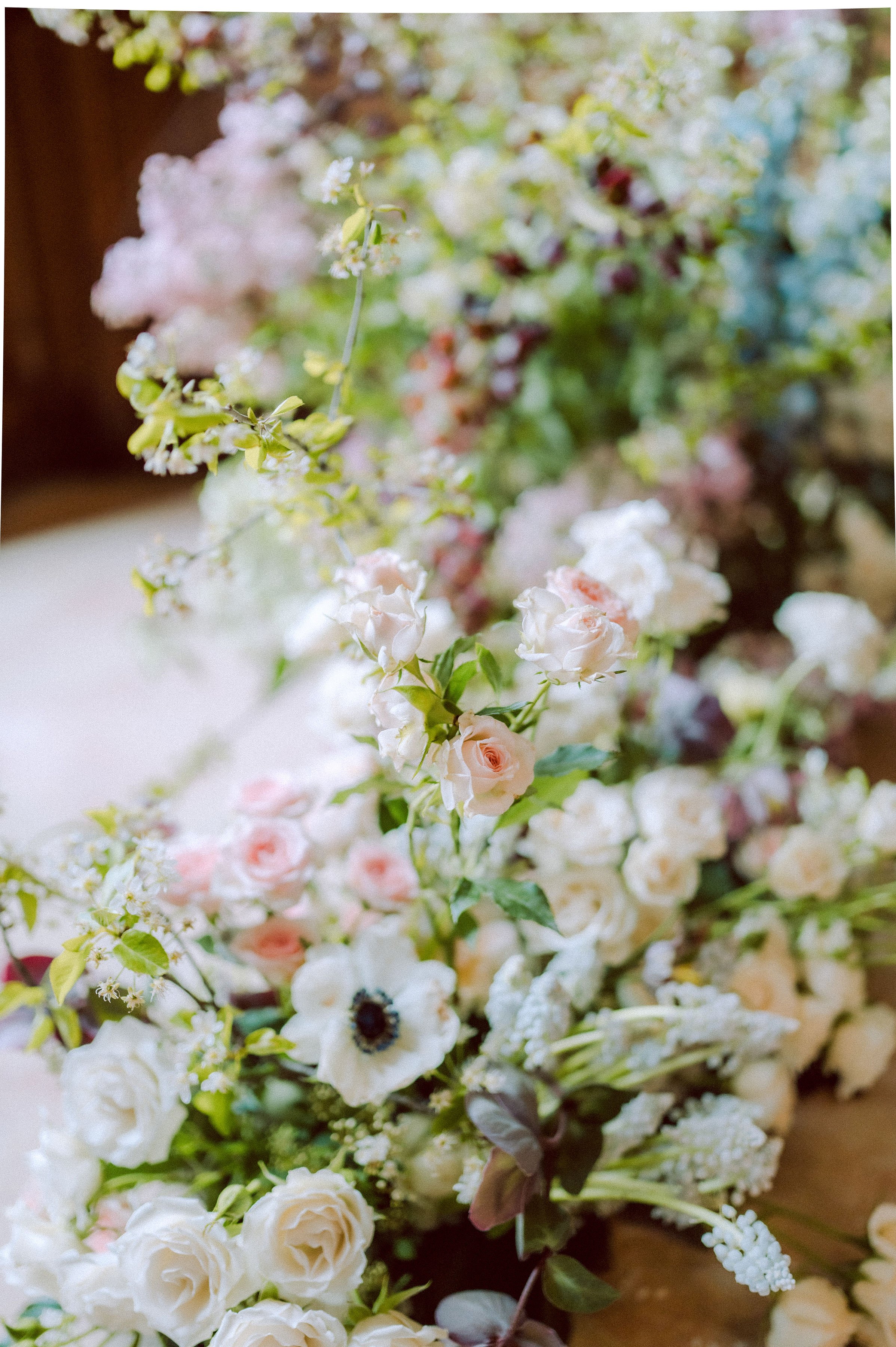 Close-up detail shot of a large, lush floral arrangement, likely a ground-level ceremony installation or aisle runner, photographed from a low angle with a shallow depth of field. The palette is soft and romantic, featuring ivory garden roses, blush spray roses, white anemones with dark centers, white muscari, pale blue delphinium visible in the background, dusty mauve berries, and abundant fresh green foliage. The flowers are arranged in a loose, garden-style manner with stems and leaves extending naturally in multiple directions, giving an unstructured, organic aesthetic. A warm wooden floor is faintly visible beneath the arrangement, suggesting an indoor venue setting.
