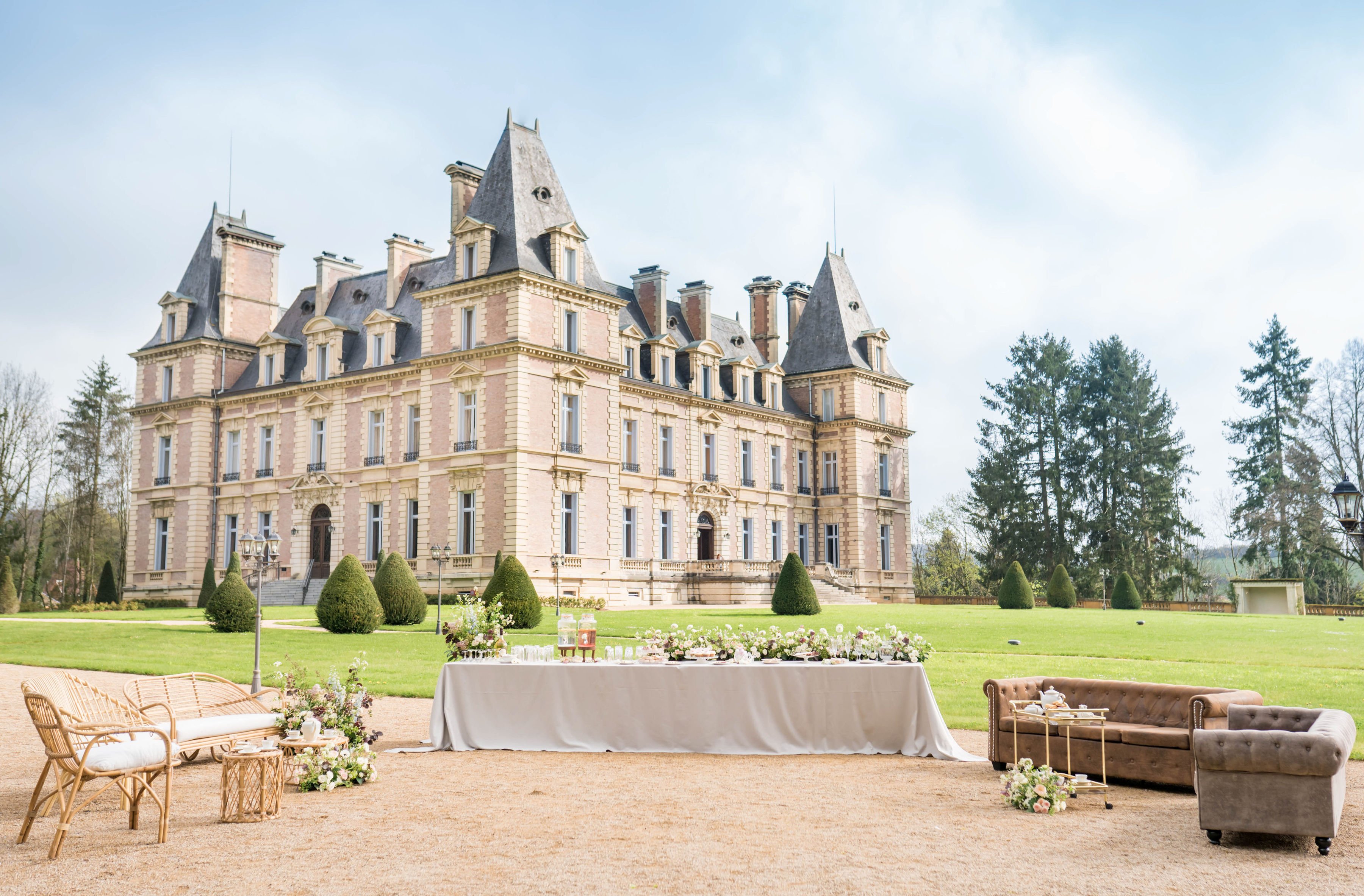An outdoor wedding cocktail hour or dessert display is set up on the gravel forecourt of a large French château, with the full facade of the multi-story pink stone building with slate-grey mansard roofs and corner turrets visible in the background. The setup includes a long rectangular table draped in a grey-taupe linen cloth, topped with florals in blush, mauve, and white tones along with glassware and small food or dessert items. Flanking the table on the left is a natural rattan loveseat with a white cushion and a small rattan side table, while on the right sits a tufted brown leather Chesterfield-style sofa with a gold bar cart beside it; both seating areas have low floral arrangements at their bases in the same blush and mauve palette. The overall styling mixes boho rattan elements with classic upholstered furniture, creating an eclectic yet cohesive outdoor lounge aesthetic. This is a wide establishing shot taken from ground level, capturing both the event décor and the full scale of the château. Potential venue feature image.