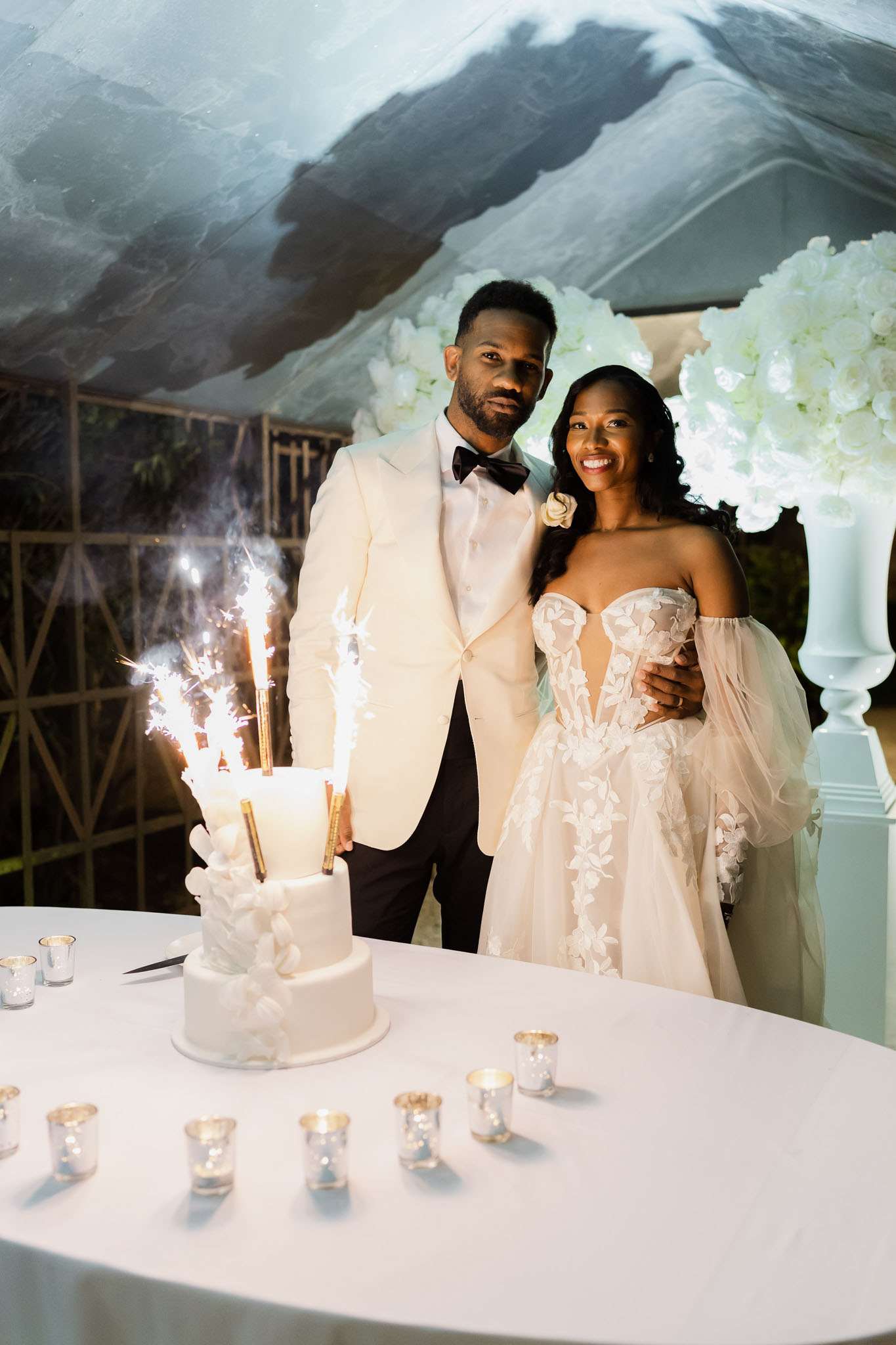 The couple poses together during the cake cutting moment at what appears to be an evening outdoor reception held under a clear-roof tent or canopy. The three-tier white wedding cake, decorated with white floral appliqués, is topped with lit sparkler candles that are actively sparkling and smoking. The table is covered in a white linen and lined with small silver mercury glass votive candle holders. The groom wears an ivory dinner jacket with black trousers and a dark bow tie, with a white floral boutonnière; the bride wears a strapless sweetheart ballgown with 3D lace floral appliqués and detachable off-the-shoulder tulle sleeves in ivory. Behind them, large white floral arrangements — appearing to be white hydrangeas and roses — are displayed on tall white pedestal urns, and a decorative gold geometric screen is visible to the left. The overall decor palette is all-white and gold with a formal, modern classic style. Medium portrait shot framing both the couple and the cake table in full.