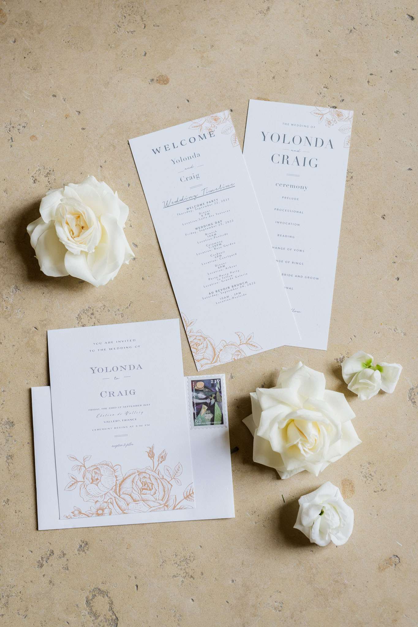 A flat lay detail shot of a wedding stationery suite for Yolonda and Craig, arranged on a warm beige stone surface. The suite includes a square invitation card, a wedding timeline card, and a tall narrow ceremony program, all printed on white paper with dark grey serif typography and copper-toned botanical line-art illustrations of roses. Scattered around the stationery are several loose ivory garden roses and small white lisianthus blooms, along with an envelope bearing two decorative postage stamps. The overall palette is white, copper, and charcoal, giving the suite a classic, refined look.