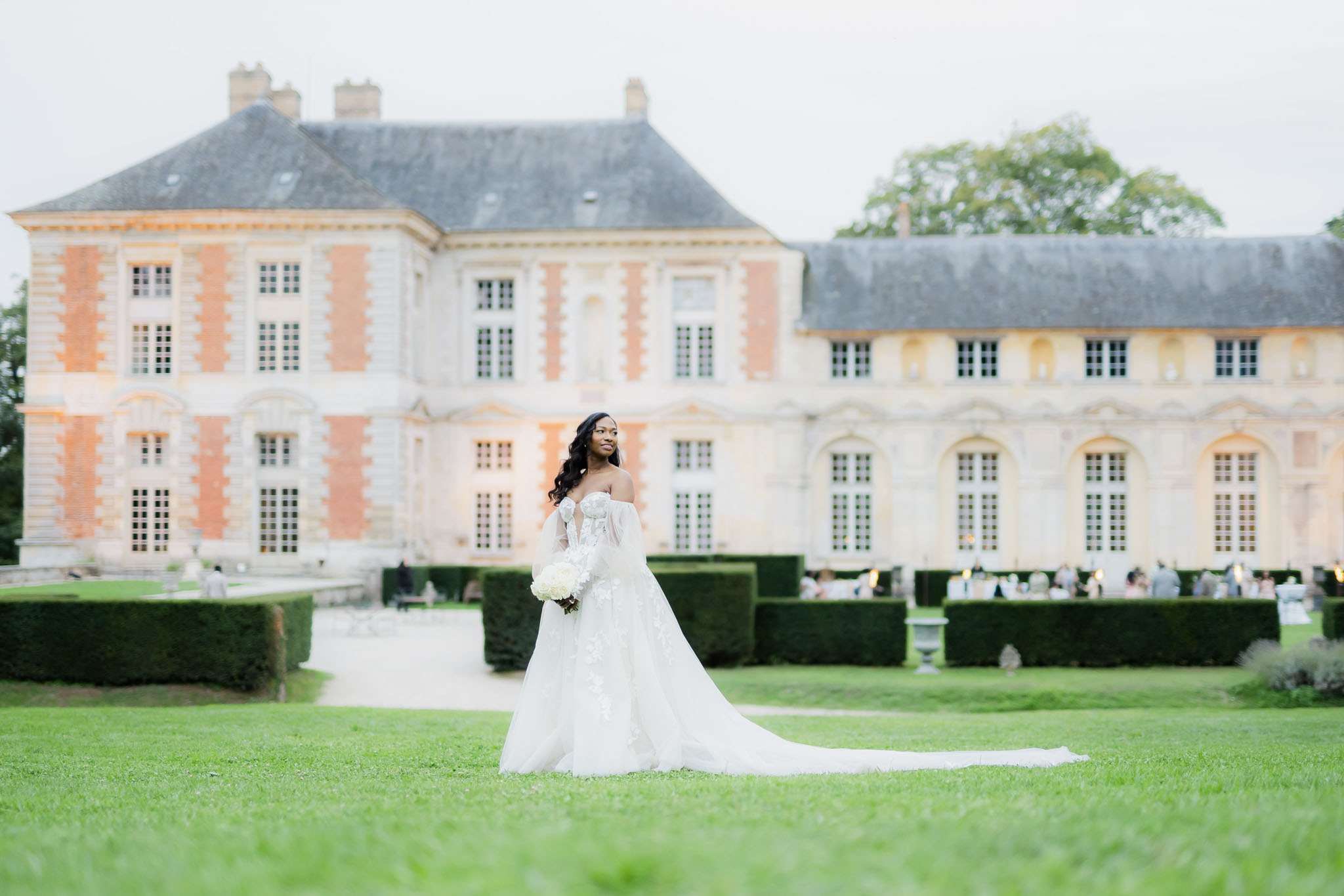A bridal portrait taken outdoors on the manicured grounds of a French chateau, with the bride standing alone on a wide lawn in front of the building. The bride wears an off-the-shoulder ball gown with lace appliqué detailing, sheer long sleeves, and an extended cathedral train spread across the grass; she holds a rounded bouquet of white peonies or hydrangeas. Her long dark hair falls in loose waves over one shoulder. In the background, the chateau features classic French architecture with warm terracotta-toned brick and stone detailing across three stories; formally clipped boxwood hedges line the garden paths, and a group of approximately 20–30 guests can be seen seated at tables in the far right background, suggesting a cocktail hour or outdoor reception in progress. The shot is a full-length wide portrait with the chateau as a backdrop. Potential venue feature image.