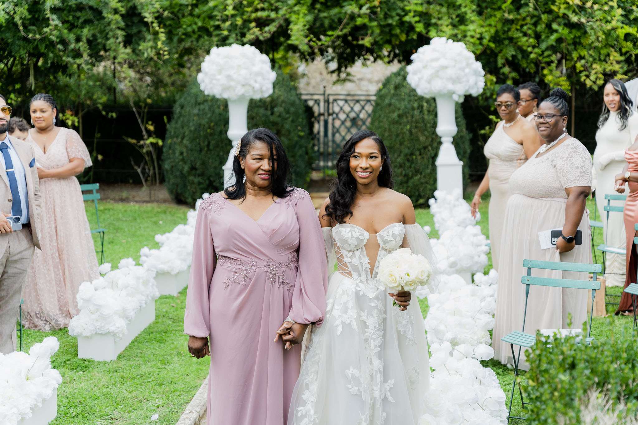 The bride is being escorted down the aisle by an older woman, likely her mother, during an outdoor garden ceremony. The bride wears an off-the-shoulder ivory gown with floral appliqué detailing and a deep V corset bodice, carrying a round bouquet of white roses and peonies. The aisle is lined with low white cube arrangements of white blooms and flanked by tall white pedestal urns topped with large white floral balls, creating an all-white floral decor scheme. Guests seated on teal bistro chairs on either side are dressed in champagne, taupe, and blush tones, while the escorting woman wears a dusty mauve long-sleeve gown with floral embroidery at the shoulders and waist. The shot is a medium wide portrait taken from ground level looking down the aisle toward the couple.