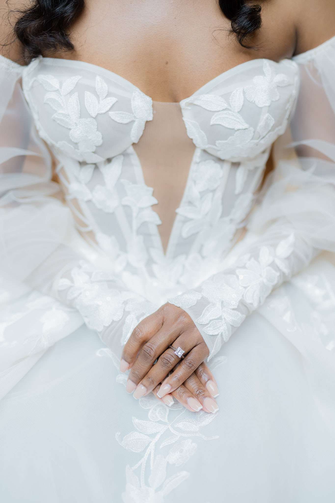 A close-up detail portrait of a bride from the chin to the lap, highlighting her white ball gown with a sweetheart neckline, structured corset bodice, and long sheer sleeves, all covered in three-dimensional appliquéd leaf and floral embroidery. Her hands are gently folded in her lap, displaying a princess-cut diamond solitaire engagement ring and a French manicure with square-shaped nails. The skirt of the gown is a smooth, voluminous white satin, contrasting with the textured lace sleeves and bodice.