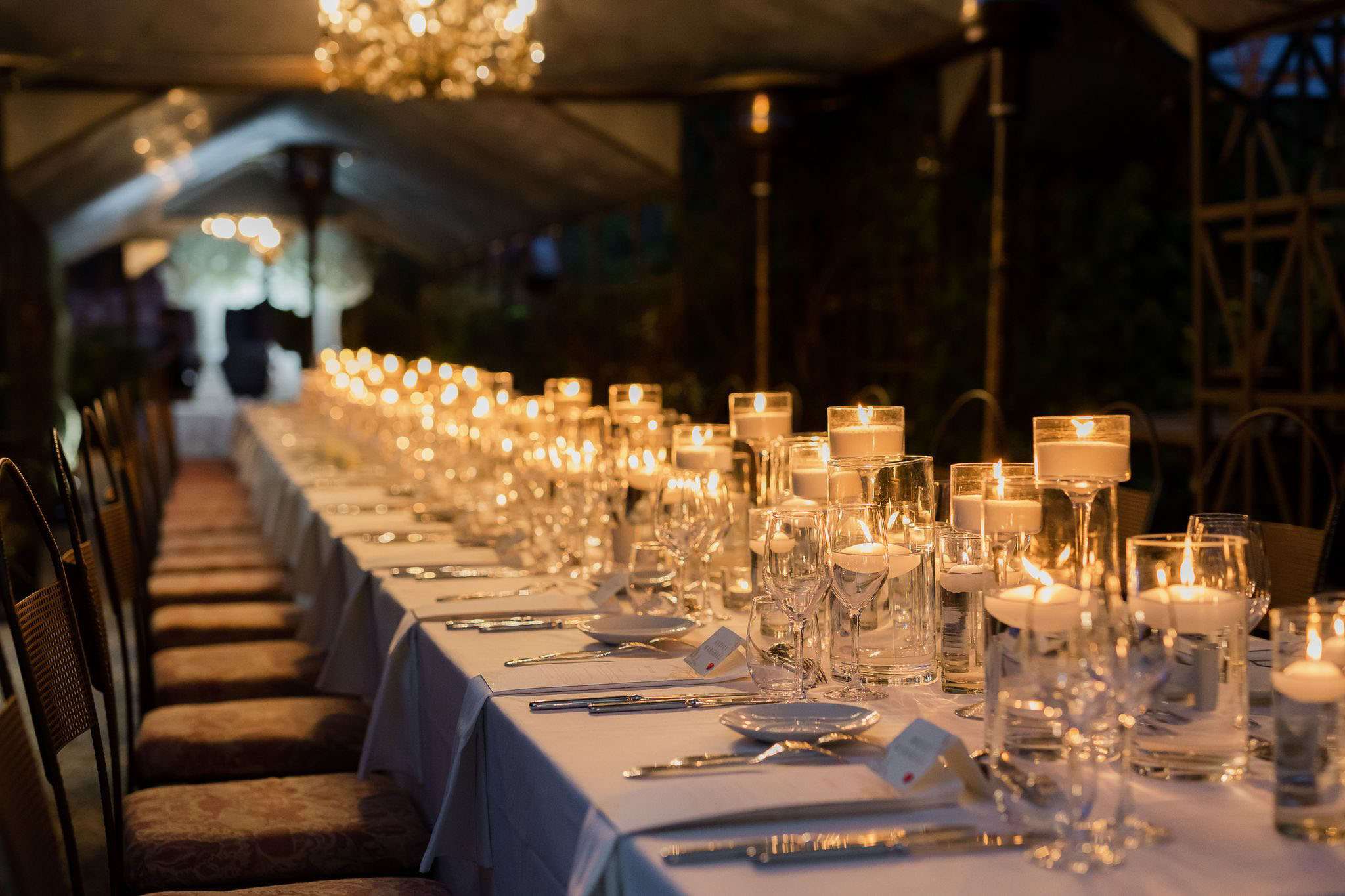 A detail shot of a long reception dinner table set up under an open-sided tent or marquee in the evening. The white-linen table is laid with silver cutlery, clear crystal wine glasses, white place cards, and silver charger plates, with rattan-style chairs lined along one side. The centerpiece decor consists entirely of clusters of floating and pillar candles in varying-height clear glass cylinders, creating a warm amber glow that runs the full length of the table. A crystal chandelier is visible overhead, and patio heaters are positioned in the background, suggesting an outdoor or semi-outdoor setting styled in a classic, candlelit aesthetic.