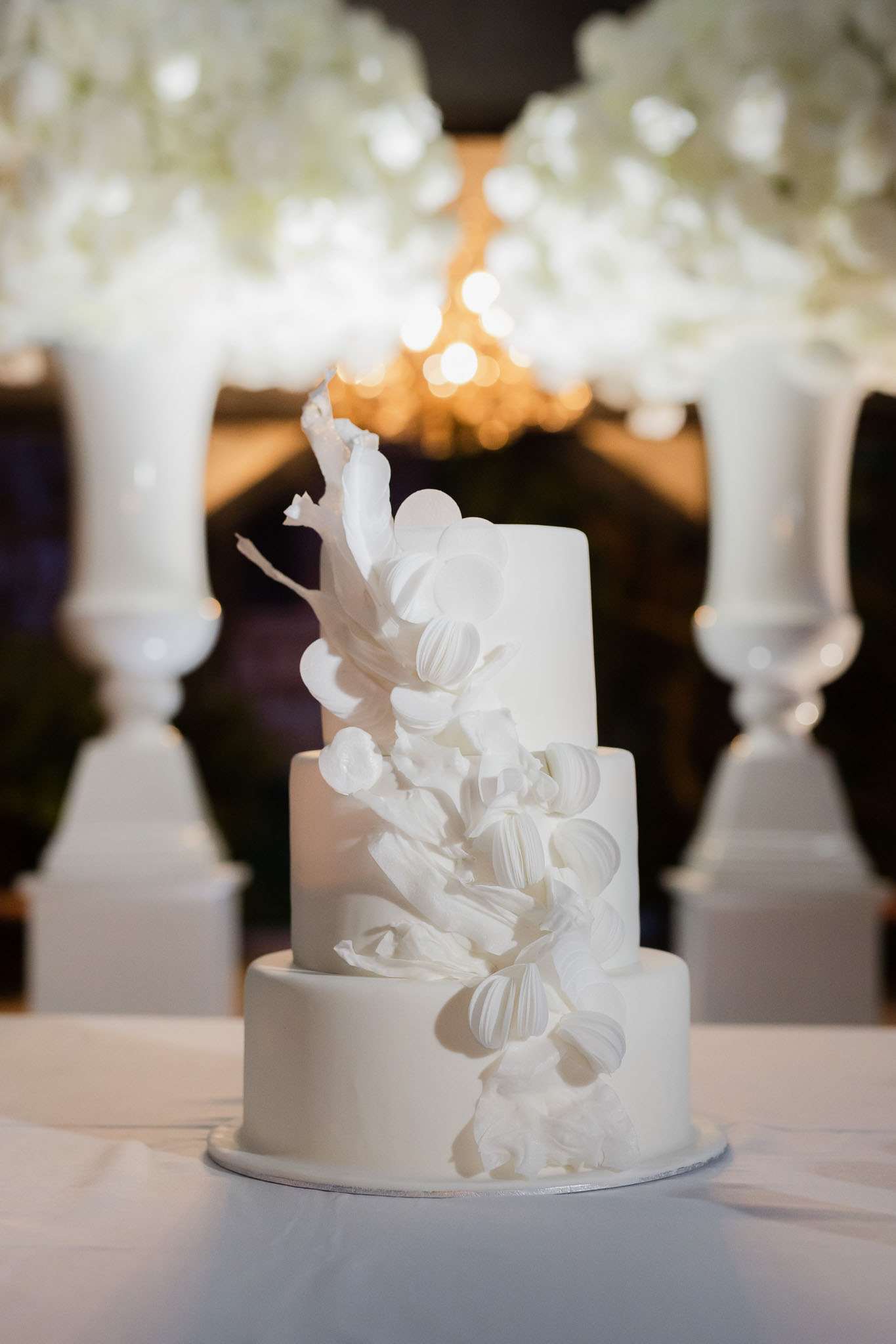 Close-up detail shot of a three-tier wedding cake covered in smooth white fondant, decorated with a cascading arrangement of white sugar flowers and ruffled petals running diagonally across all three tiers. The cake sits on a white linen-covered table in what appears to be an indoor reception venue. In the soft-focus background, two tall white urn-style pedestals flank the cake, topped with large white floral arrangements — likely white hydrangeas — and warm chandelier lighting creates golden bokeh behind the cake. The overall decor palette is entirely white and gold, consistent with a classic, all-white monochromatic reception styling.