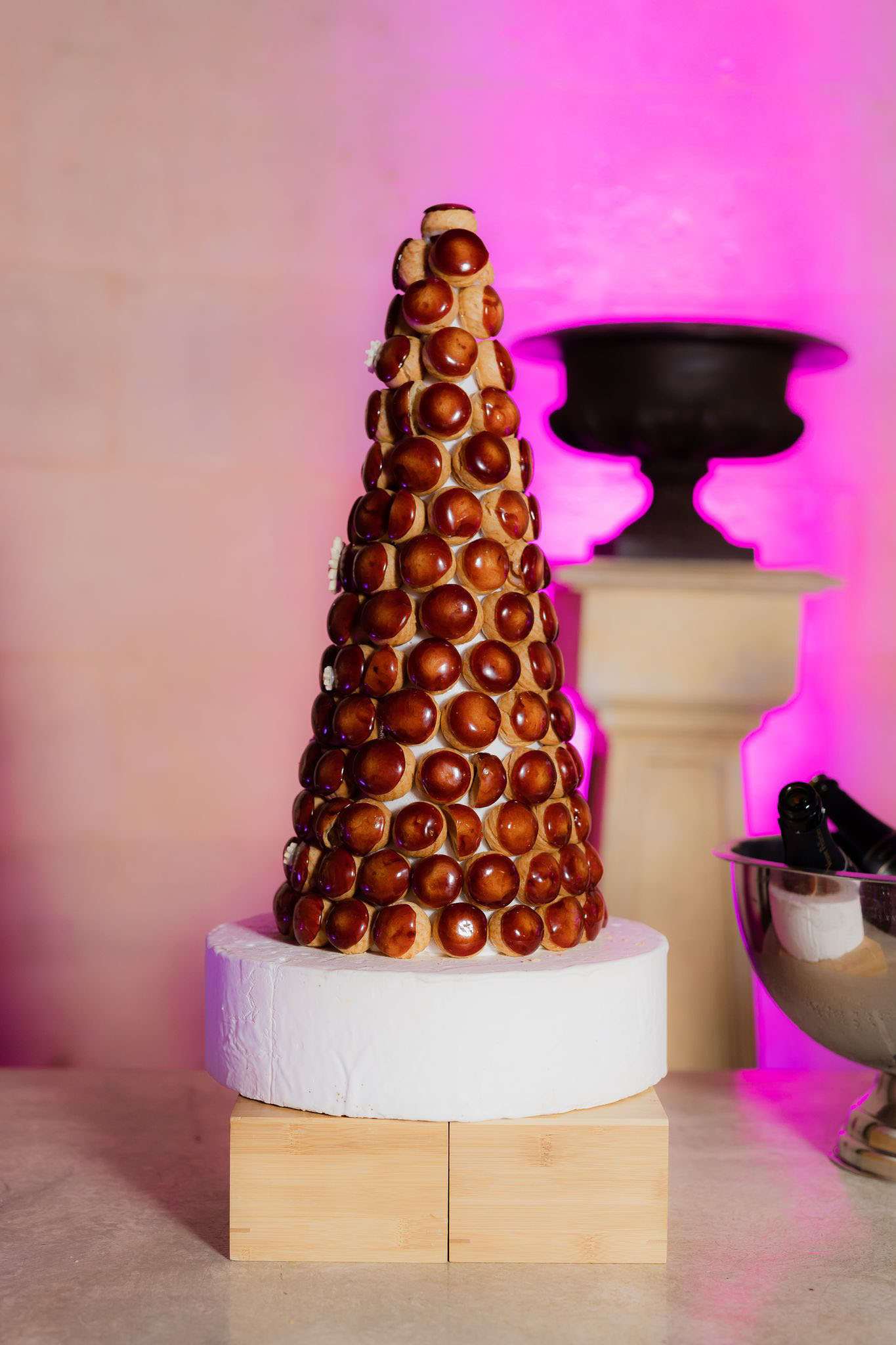 Close-up detail shot of a traditional French wedding croquembouche — a tall conical tower of caramel-glazed choux puffs — displayed on a white nougat or sugar base set on two small bamboo wooden blocks. The profiteroles are a deep amber-brown from the caramel coating, with small white sugar flower accents visible at intervals along the tower. The dessert is placed on a marble-effect surface, with a silver champagne ice bucket partially visible to the right and a stone pedestal bearing a dark decorative urn in the background. The backdrop wall is lit with a vivid magenta/hot pink wash of uplighting, creating a bold, festive atmosphere.