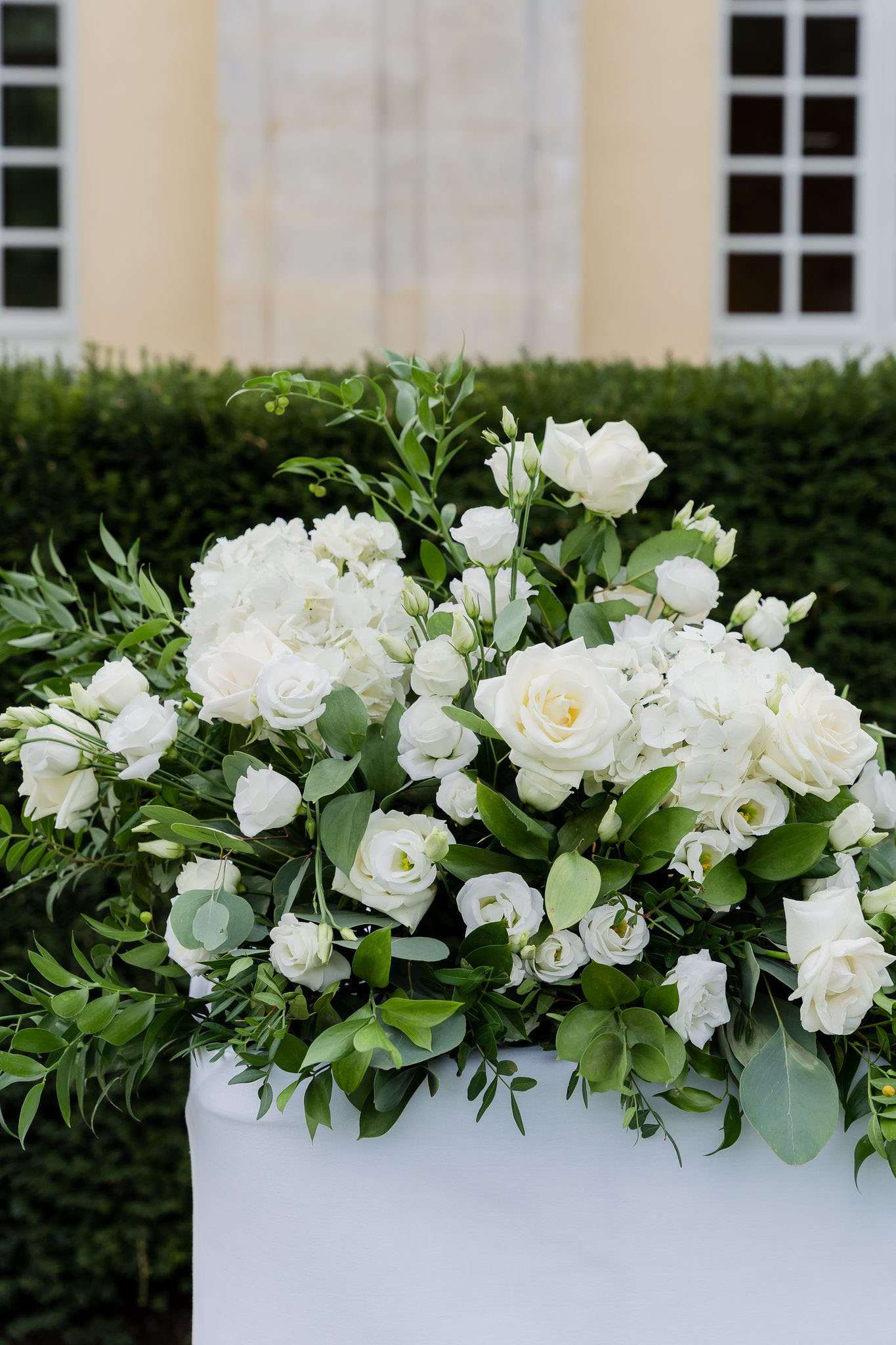 A close-up detail shot of a large floral arrangement placed on top of a white-draped pedestal or ceremony altar, photographed outdoors. The arrangement features white garden roses, white hydrangeas, white lisianthus, and abundant mixed greenery including eucalyptus and olive branches, styled in a lush, overflowing design. The decor palette is strictly white and green, consistent with a classic, clean aesthetic. In the background, a neatly trimmed hedge and the pale yellow facade of what appears to be a chateau or formal venue building with white-framed windows are softly out of focus.
