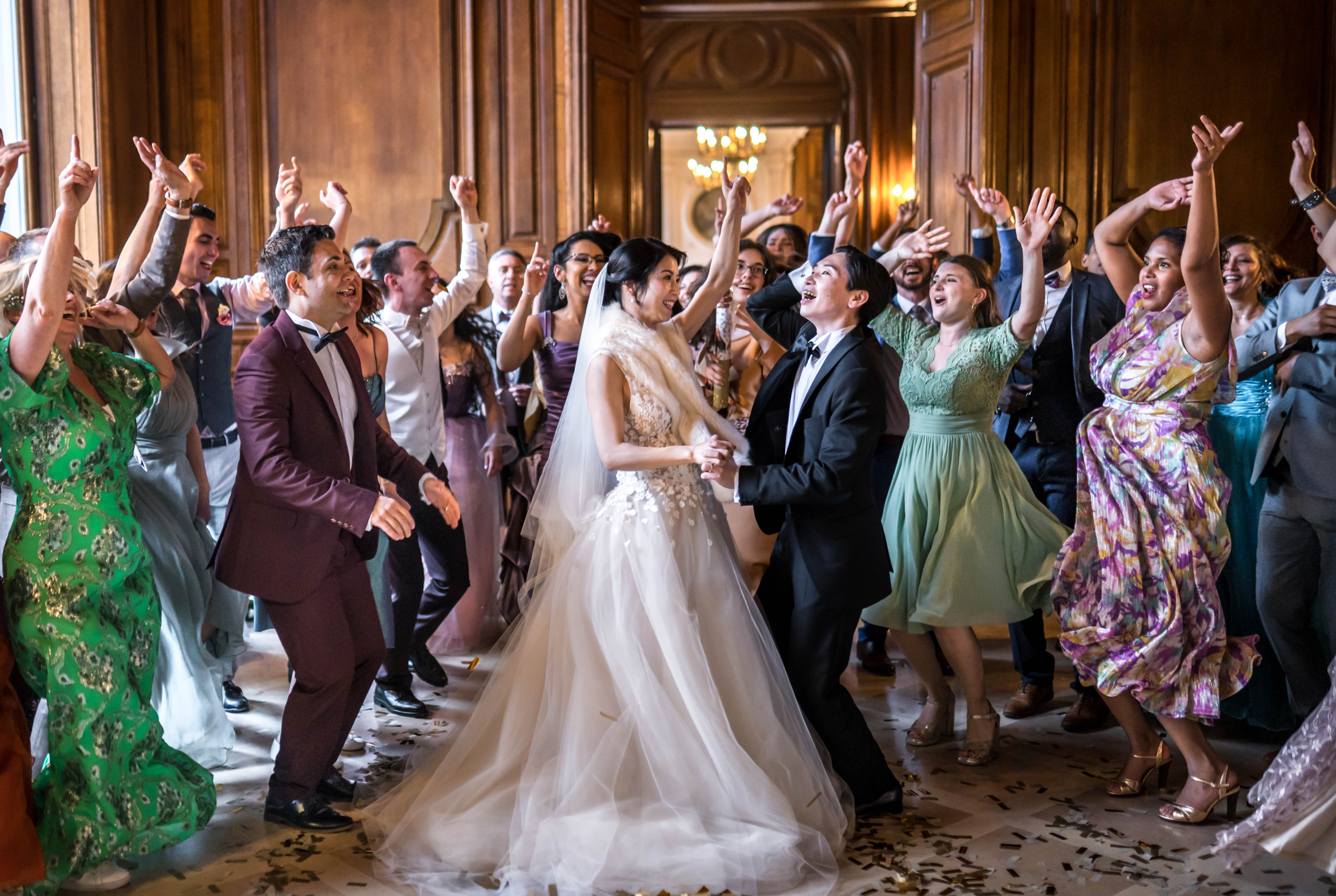 The couple shares a celebratory first dance surrounded by approximately 30 guests inside a formal ballroom with floor-to-ceiling carved wood paneling and a crystal chandelier visible in the background. The bride wears a voluminous ivory tulle ball gown with floral appliqué bodice, a long cathedral veil, and a cream feather or fur wrap over her shoulders, while the groom wears a classic black tuxedo with bow tie. Guests are dancing energetically with arms raised, dressed in a mix of colorful formal attire including a burgundy tuxedo, sage green midi dress, green and gold printed gown, and a multicolored floral dress, with gold confetti scattered across the marble floor. The scene has a high-energy, celebratory atmosphere with warm ambient lighting from the chandeliers, captured in a wide environmental shot that shows the full sweep of the room and the crowd surrounding the couple.