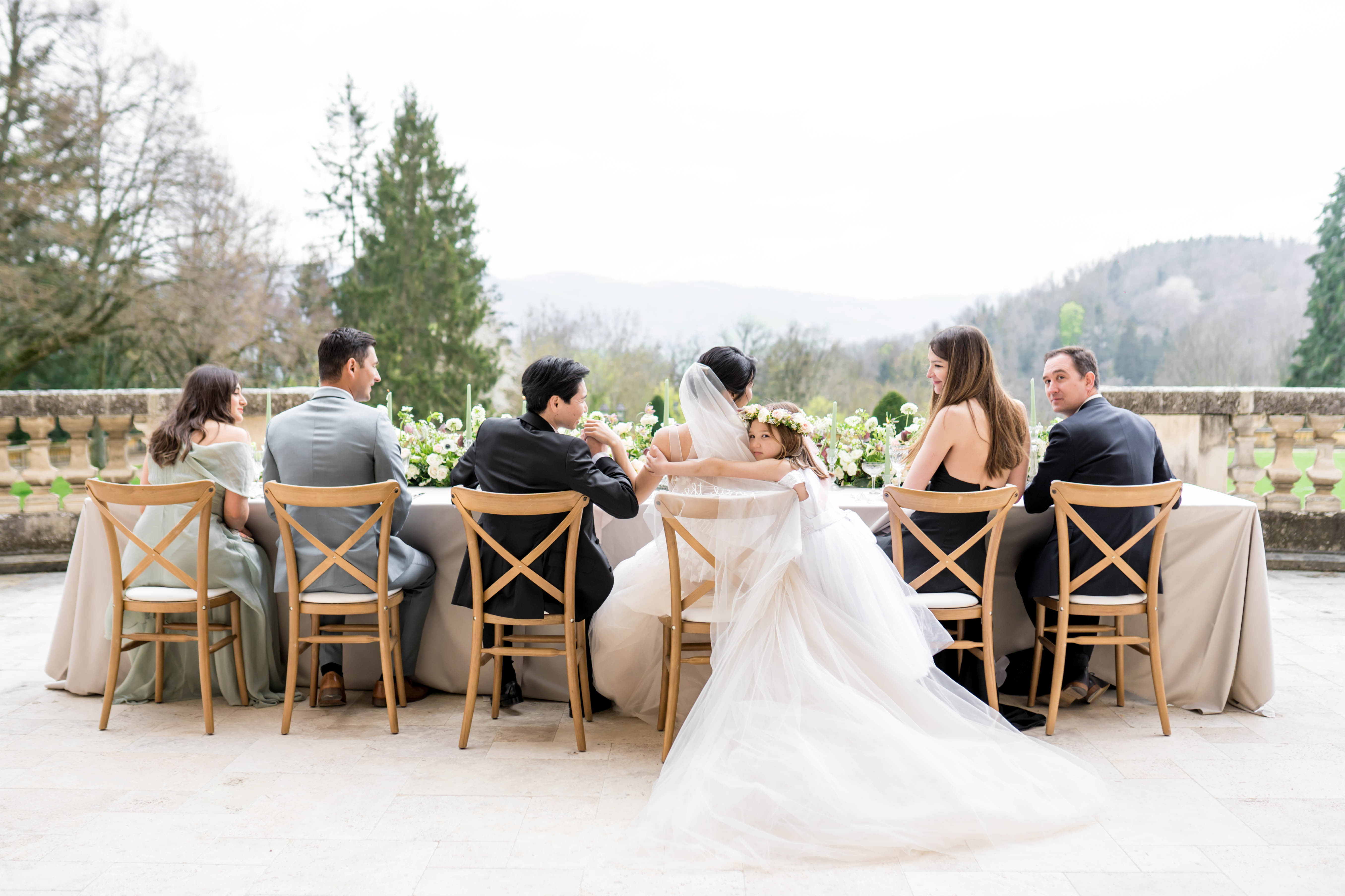 A wedding reception dinner scene set outdoors on a stone terrace with a balustrade railing, overlooking a wooded hillside landscape. Seven guests and the couple are seated at a rectangular table draped in a taupe linen, with natural wood cross-back chairs; the bride sits at center with her back partially to the camera, wearing a white ballgown with a long veil and a floral crown of blush and ivory blooms, while the groom beside her wears a black suit. The table centerpiece features a lush low arrangement of white and blush flowers — including what appear to be ranunculus and anemones — with trailing greenery and tall tapered sage-green candles. Guests are dressed in a coordinated palette of sage green off-the-shoulder gown, grey suit, black tuxedo, black halter dress, and dark navy suit, giving the styling a classic, polished feel. The wide-angle shot is taken from behind the table, capturing the full spread of the bride's gown train pooling on the stone floor in the foreground.