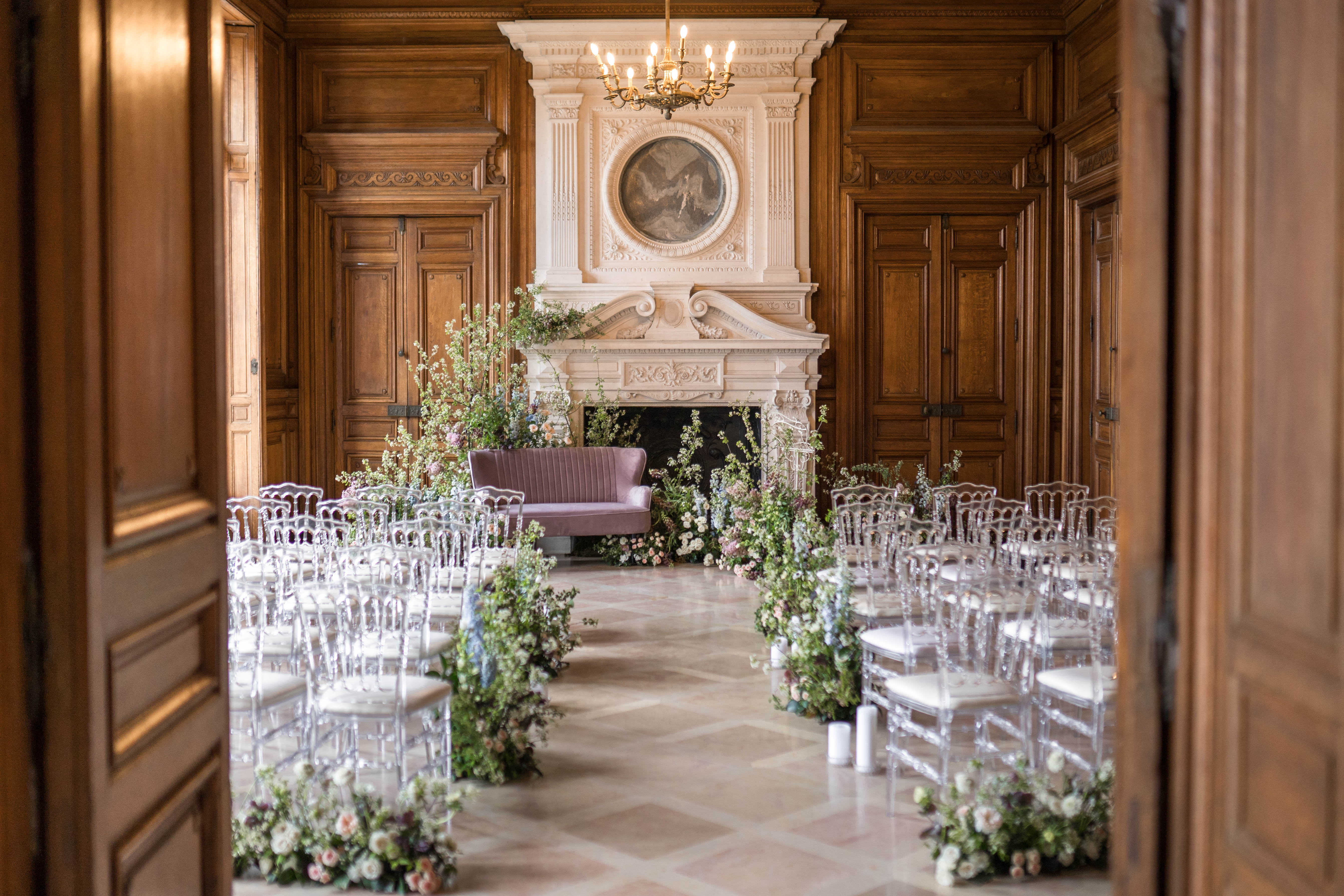 An indoor ceremony space set up inside a French chateau-style room, photographed from the doorway in a wide shot framed by tall wooden paneled doors. The room features floor-to-ceiling warm walnut boiserie paneling, a grand carved white stone fireplace with an oval painted medallion above it, and a brass chandelier overhead. Clear acrylic Chiavari chairs with ivory cushions are arranged in two rows along a central aisle, with clusters of lush floral arrangements placed at the base of each row — composed of blush and mauve roses, white blossom branches, soft blue florals, and trailing greenery. The ceremony focal point is a dusty mauve velvet loveseat positioned in front of the fireplace, flanked by two large asymmetrical floral installations of the same palette with tall branching stems. White pillar candles are placed along the aisle at floor level. The overall decor palette is soft blush, mauve, and sage green with a classic French interior aesthetic. Potential venue feature image.