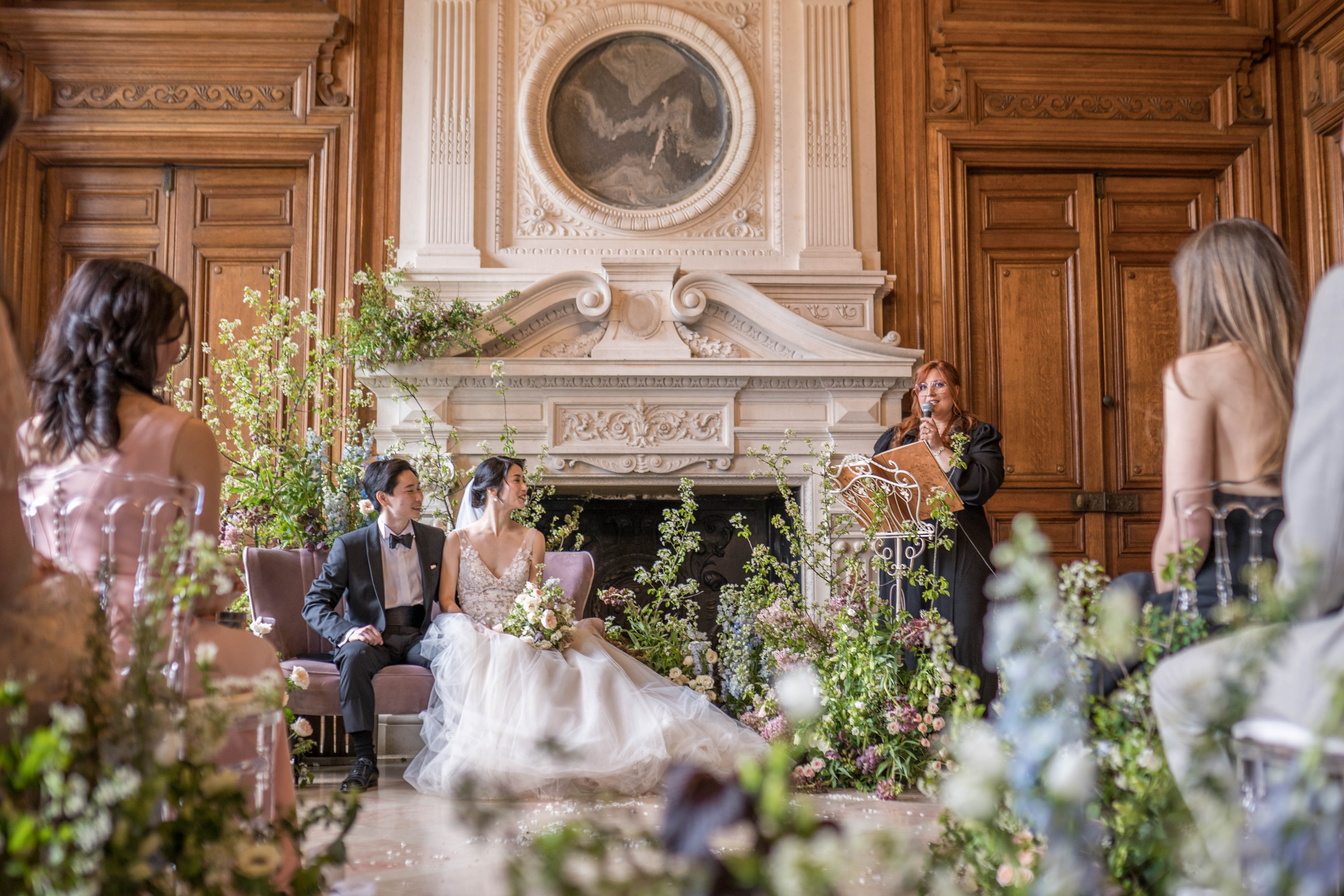An indoor wedding ceremony taking place in a grand room with ornate carved wood paneling and a large white stone fireplace as the focal backdrop. The couple is seated on mauve-toned chairs in front of the fireplace — the groom wearing a black tuxedo with bow tie and the bride in a tulle ball gown with a floral-appliqué bodice, holding a bouquet of blush and cream blooms — while an officiant in a black dress speaks into a microphone at an ornate white iron lectern. The ceremony decor features extensive garden-style floral installations composed of tall branching white flowering shrubs, lush greenery, blush roses, and soft lavender florals arranged loosely around the fireplace and along the floor, creating a naturalistic, garden-in-bloom aesthetic. The shot is taken from a low angle looking through foreground floral arrangements toward the couple, with guests partially visible on either side, giving a wide, immersive perspective of the ceremony setting.