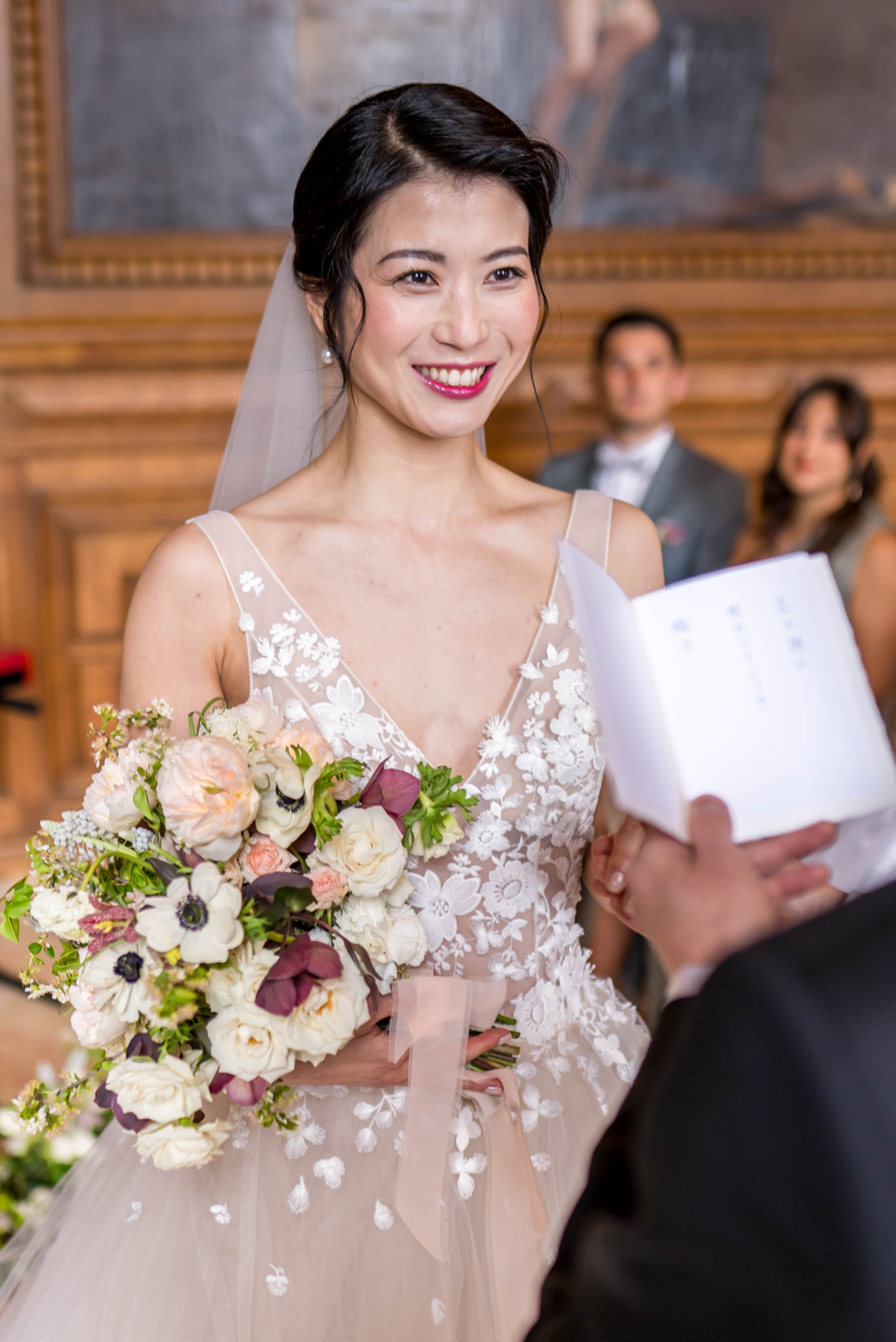 A close-up portrait of a bride during an indoor ceremony, smiling warmly toward the officiant who holds an open ceremony booklet in the foreground. She wears a blush tulle ballgown with a sheer V-neckline bodice featuring white floral appliqué, paired with a plain veil, pearl earrings, and a bold pink-red lip. Her bouquet is a loosely arranged mix of blush garden roses, ivory spray roses, white anemones with dark centers, deep burgundy hellebores, and green foliage, tied with a blush ribbon. The setting appears to be an ornate indoor hall with warm wood paneling and large oil paintings visible in the background, suggesting a formal civic or chateau venue. A groom in a grey suit with bow tie and a female guest are visible in the soft background.