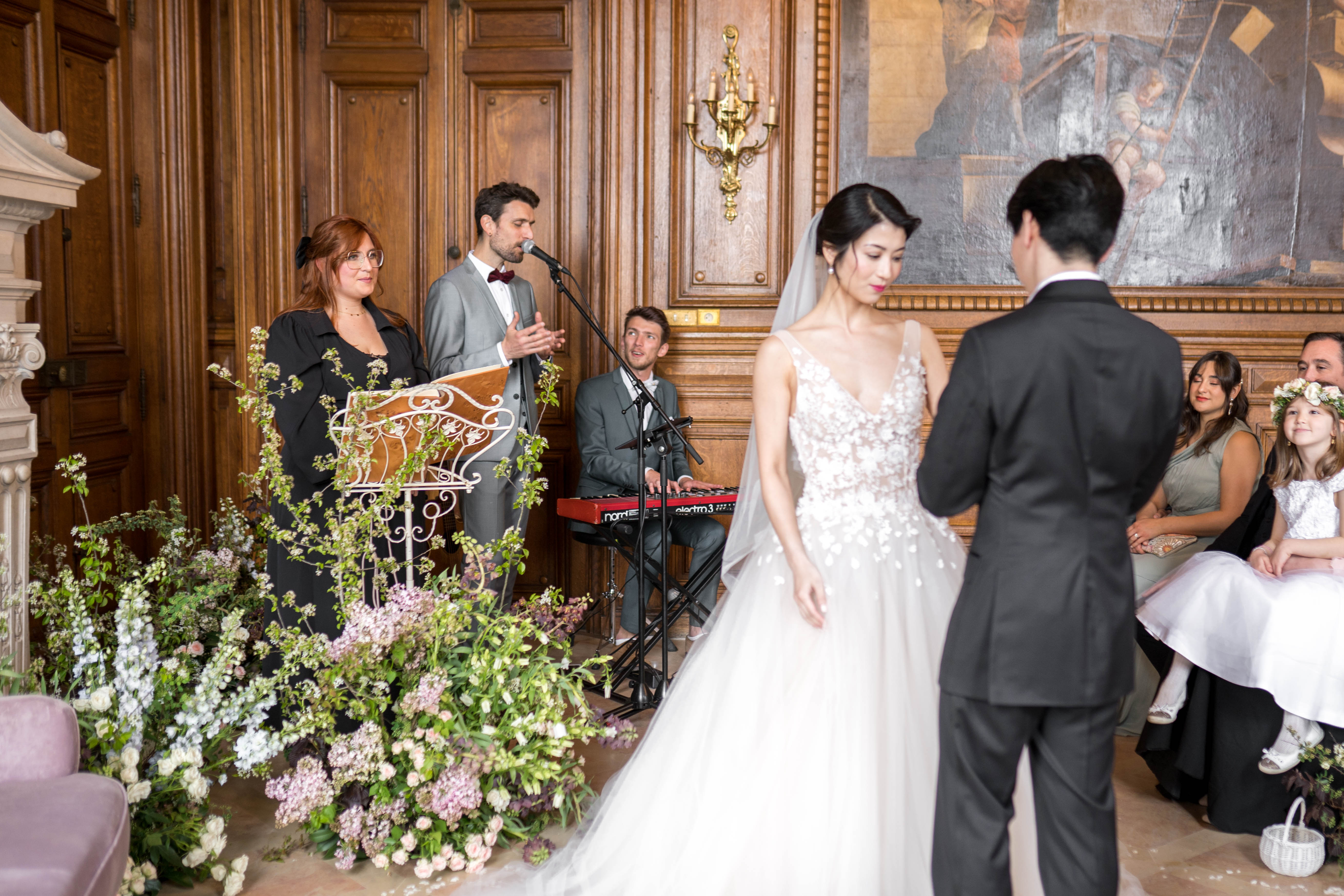 An indoor wedding ceremony taking place in a chateau-style room with dark wood paneling, a gold wall sconce, and a large classical oil painting on the wall. The bride stands in the foreground wearing a white ball gown with a heavily floral-appliquéd bodice and a long veil, facing the groom who is dressed in a dark charcoal suit; both are seen from behind or in three-quarter profile. A male singer in a grey suit with a burgundy bow tie performs at a microphone stand beside a female officiant in a black dress at an ornate white iron lectern, while a second musician plays a red Nord keyboard behind them. The ceremony altar area is decorated with an abundant ground-level floral installation featuring white, blush pink, and pale lavender blooms with cascading greenery and branches. Seated guests visible on the right include a woman in a sage green dress and a young flower girl in white holding a small white basket.