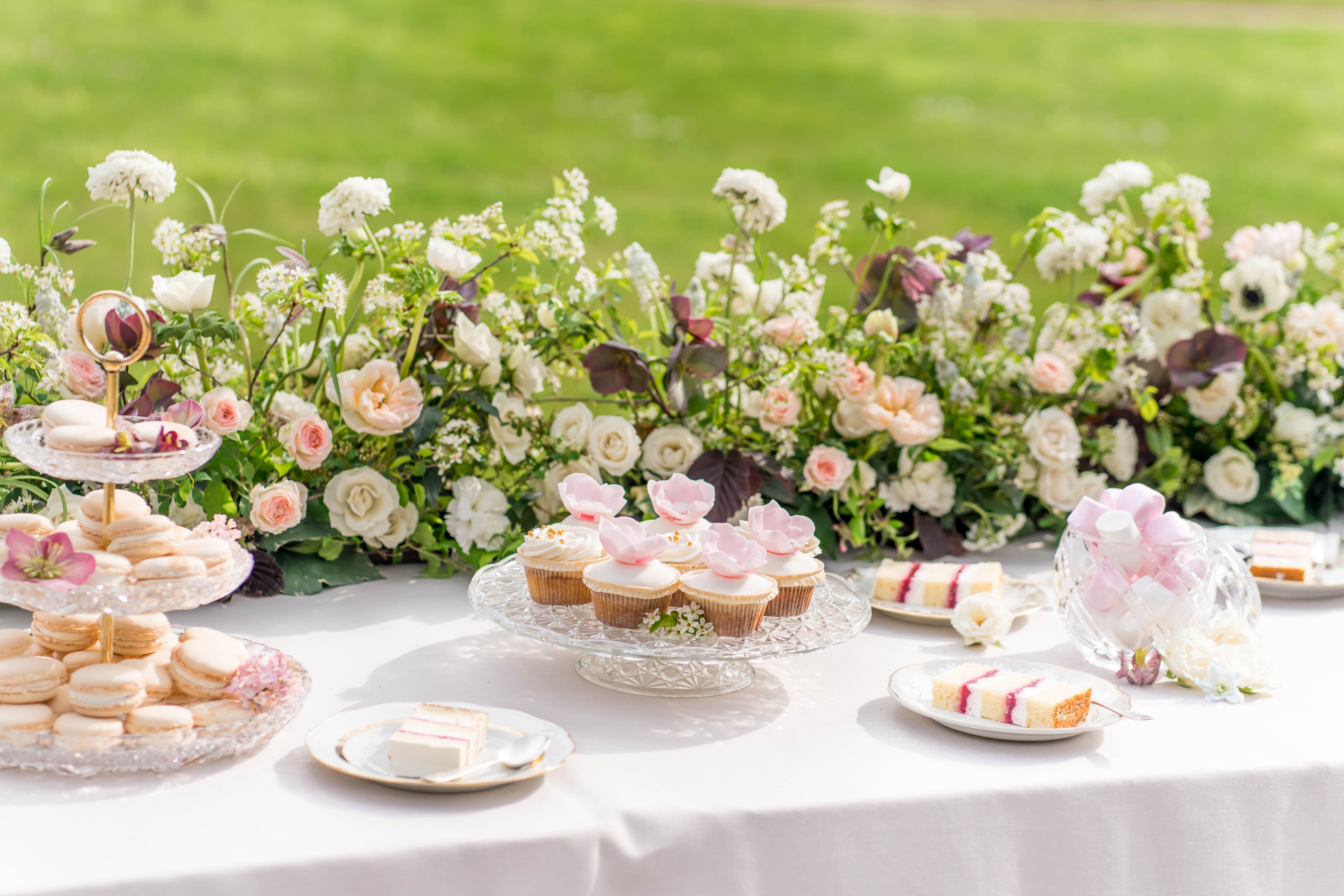 An outdoor wedding dessert table covered in a white linen, photographed in a wide close-up shot. The table displays a tiered crystal stand stacked with ivory French macarons, a crystal pedestal plate holding white-frosted cupcakes topped with blush pink sugar flower petals, small white cake slices with berry jam filling on individual plates, and additional pastries on crystal serveware. A lush low floral arrangement runs along the back of the table featuring ivory garden roses, blush peach roses, white anemones, deep burgundy hellebores, and greenery with dark foliage accents, creating a romantic garden-style dessert display. The overall color palette is white, blush pink, and burgundy with gold metallic accents on the tiered stand, consistent with a classic garden wedding aesthetic.