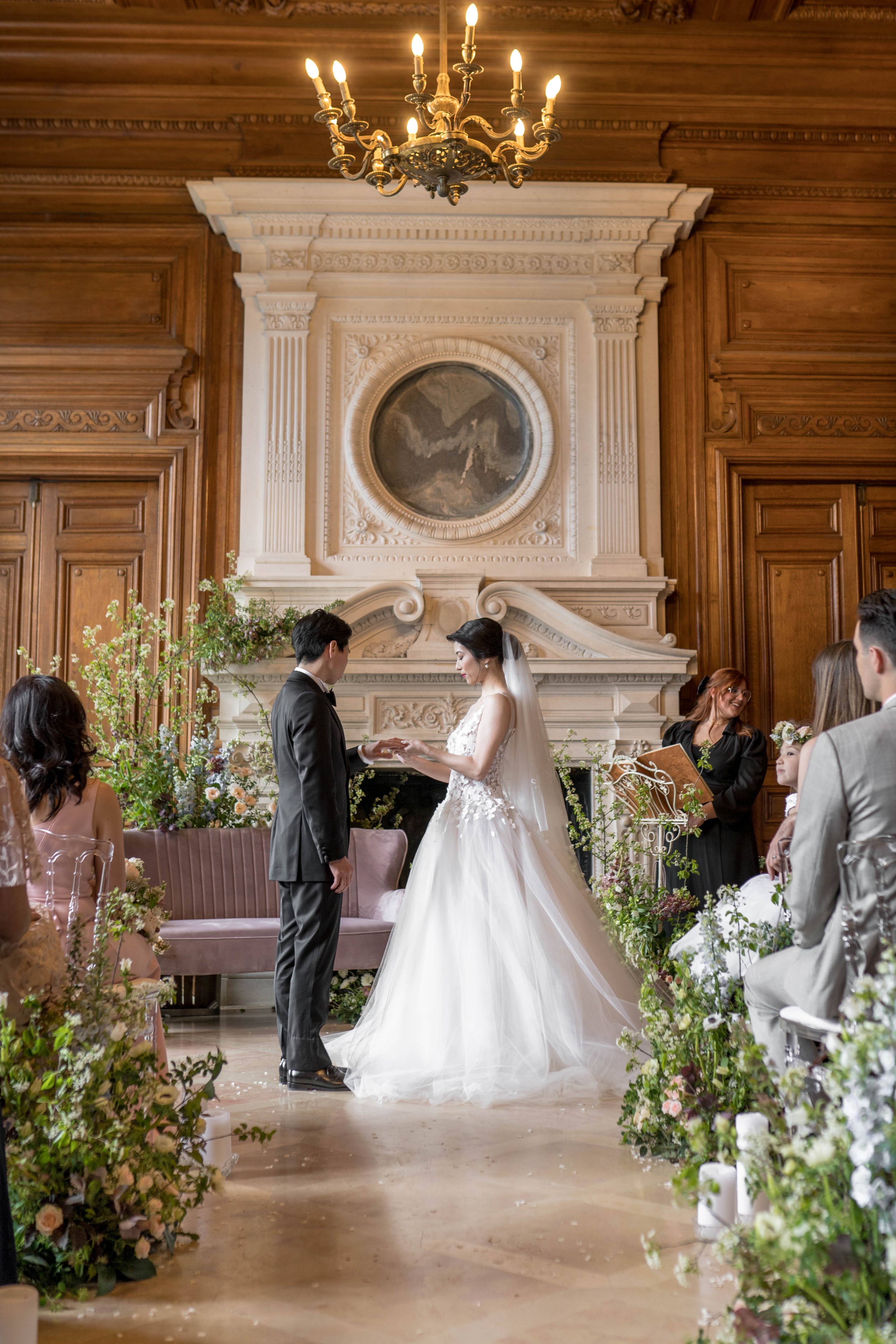 An indoor wedding ceremony taking place in a grand room featuring dark wood-paneled walls, an ornate cream stone fireplace with carved detailing and a central circular painting, and a gold candelabra chandelier overhead. The couple stands at the altar exchanging rings — the groom wears a dark charcoal suit with a bow tie, and the bride wears a white ballgown with a lace appliqué bodice and a long cathedral veil. The officiant stands behind them dressed in black, holding a wooden book. Guests are seated on either side on acrylic and upholstered chairs, with a dusty mauve velvet sofa visible behind the couple. The aisle and altar area are flanked by abundant loose garden-style floral arrangements featuring soft peach and blush blooms, white flowers, and generous amounts of trailing greenery. The decor palette combines blush, cream, and green tones against the warm wood interior, giving a classic yet organic styling. Wide shot taken from the back of the aisle looking toward the fireplace. Potential venue feature image.