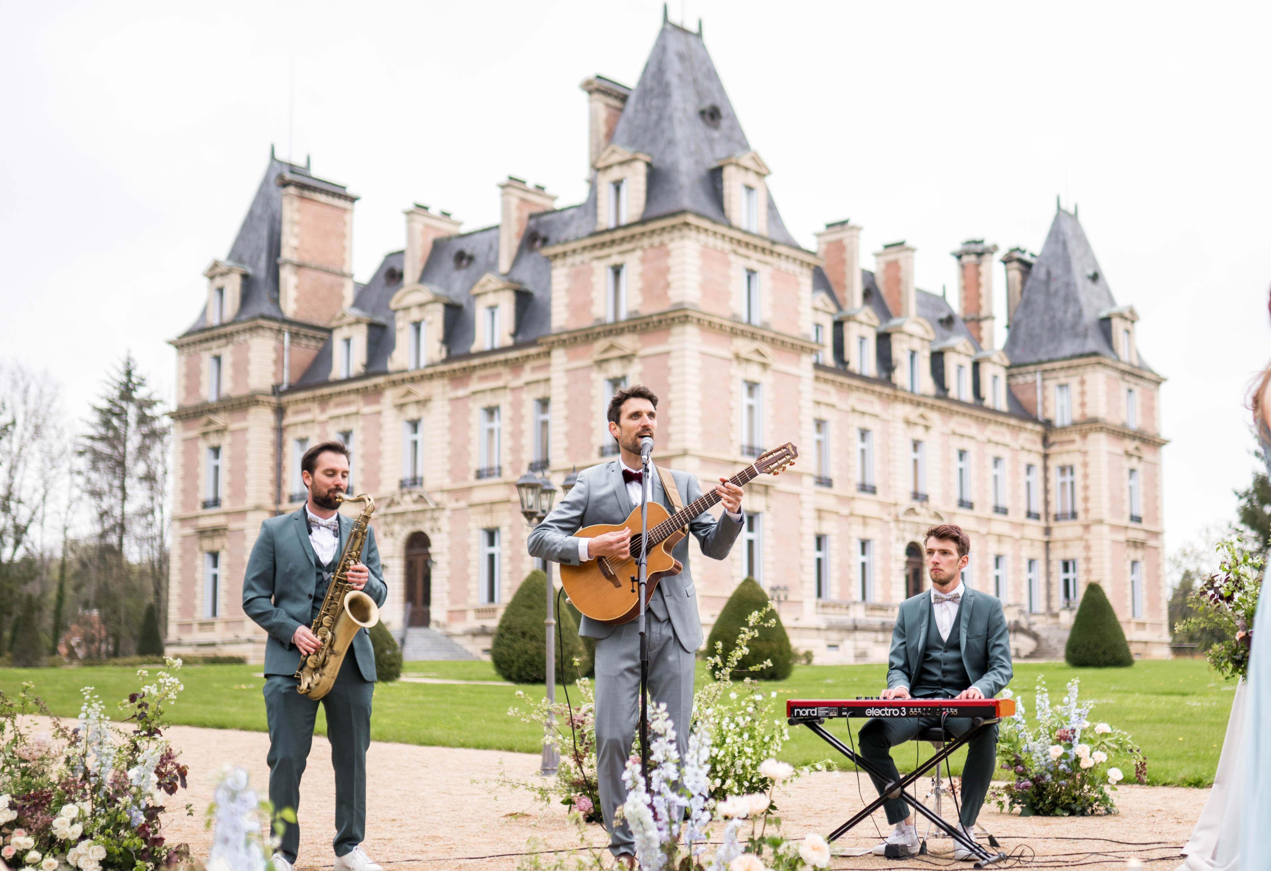 A three-piece live band performs outdoors in front of a grand French château during what appears to be a wedding ceremony or cocktail hour. The center musician plays an acoustic guitar and sings into a microphone, flanked by a saxophonist on the left and a keyboardist playing a Nord Electro 3 on the right; all three wear matching teal-green suits with bow ties, with the guitarist in a light grey suit and burgundy bow tie. The foreground features low floral arrangements in a palette of blush, ivory, dusty blue delphiniums, and deep burgundy blooms. The wide shot places the performers centrally with the multi-story pink-brick château with dark slate mansard roofs as the backdrop. Potential venue feature image.