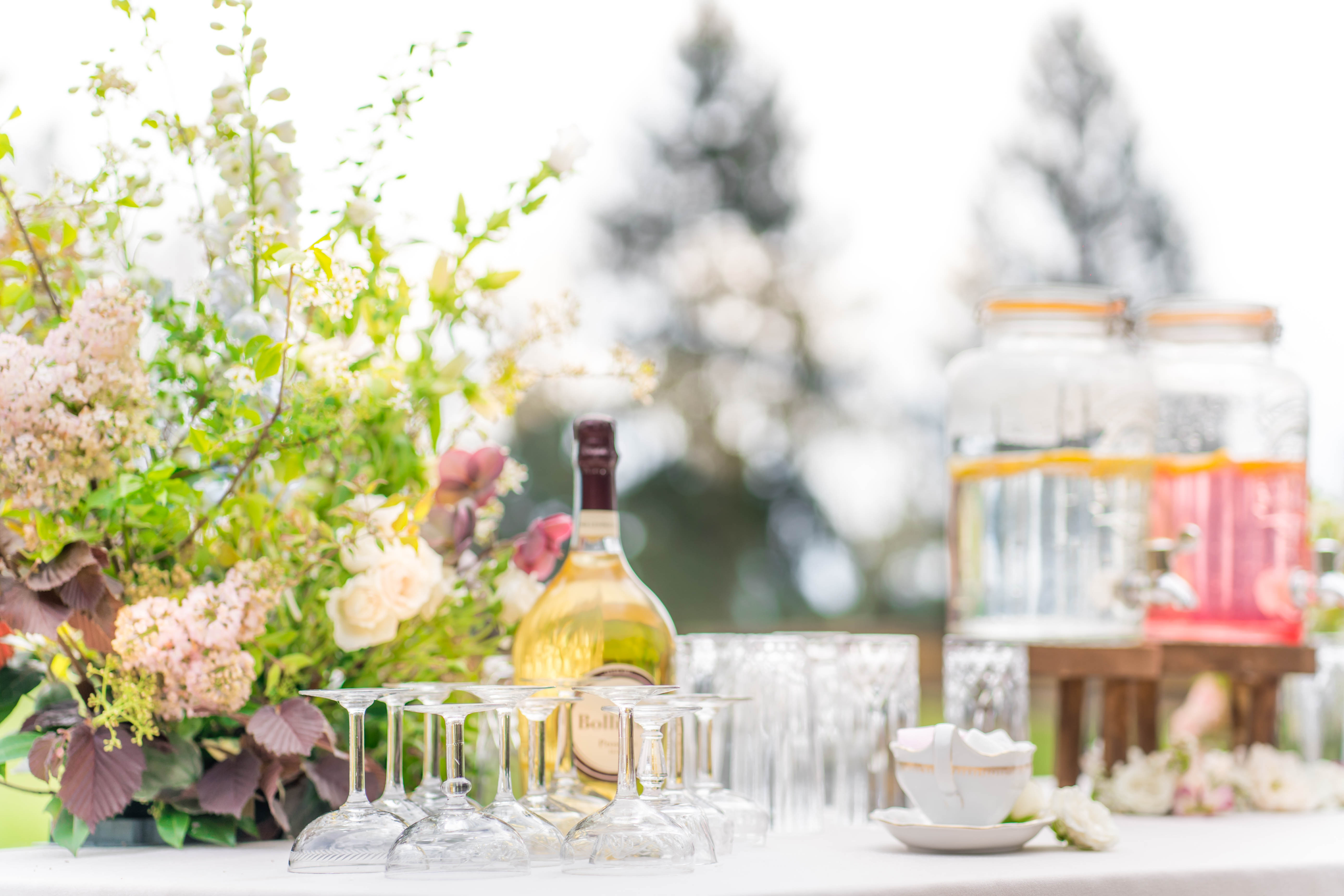 Close-up detail shot of an outdoor cocktail hour bar setup, with no people visible. The foreground features a row of upside-down crystal wine glasses arranged on a white linen surface alongside a bottle of Baileys liqueur and a white porcelain cup with gold rim detailing on a matching saucer. To the right in soft focus, a wooden beverage dispenser station holds multiple glass dispensers containing colorful drinks in shades of pink, red, and clear. A large floral arrangement on the left incorporates blush hydrangeas, cream garden roses, burgundy foliage, white foxglove-style blooms, and trailing greenery in a garden-style, loosely structured design. The overall decor palette is blush, cream, and burgundy with a romantic, garden-party aesthetic.