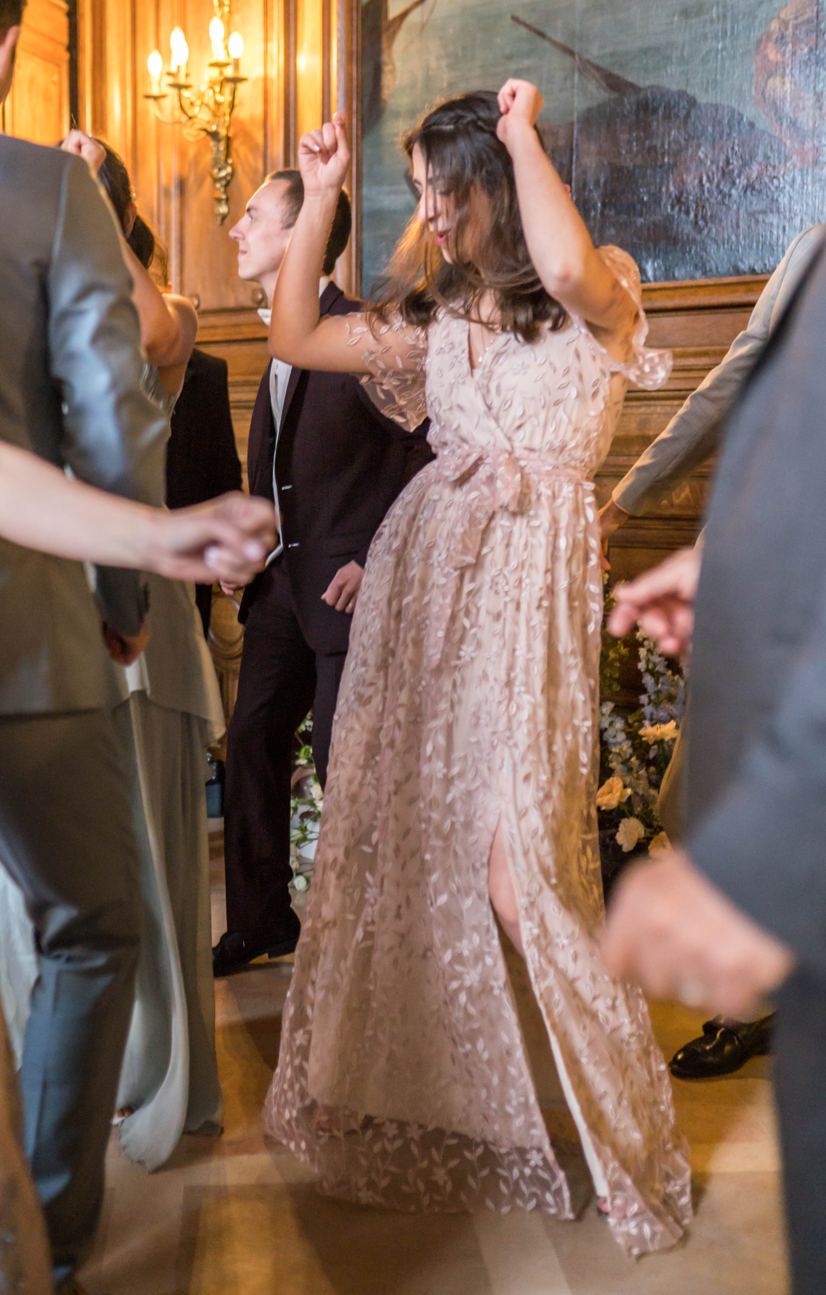 A reception dance floor scene captured indoors, likely in a chateau or manor, with wood-paneled walls, a gold candelabra wall sconce, and a large oil painting visible in the background. A woman in a floor-length blush pink lace gown with floral appliqué detailing and a front slit dances energetically with her arms raised, surrounded by several other guests in formal attire including dark tuxedos and grey suits. A floral arrangement of peach, white, and lavender blooms is visible at floor level in the background. The image is a candid mid-shot taken from within the crowd, giving it an immersive, in-the-moment feel.