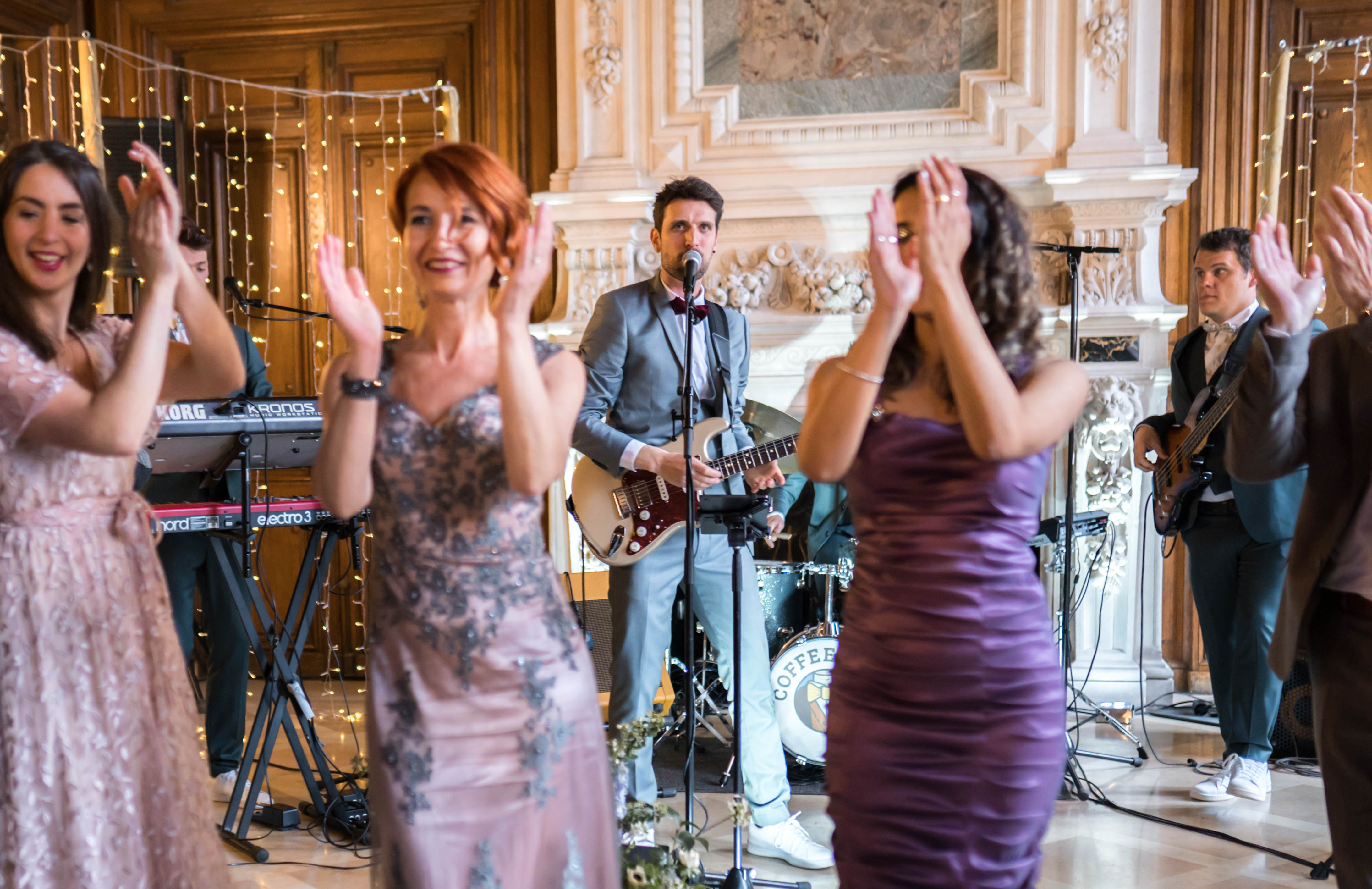 A live band performs during a wedding reception in an ornate indoor ballroom or chateau salon featuring dark wood paneling, an elaborately carved white marble fireplace, and warm fairy lights draped along the walls. The band includes a male vocalist and guitarist in a light grey suit with a burgundy bow tie, a bassist, a keyboardist playing a Korg Kronos and a Nord Electro 3, and a drummer partially visible behind a kick drum branded 'Coffee.' In the foreground, several female guests clap and dance, dressed in a blush pink lace gown with grey floral embroidery, a purple strapless ruched dress, and a pale pink embellished gown. The medium-wide shot captures both the performing musicians and the energetic response of approximately four to five guests in the foreground, with the reception's classic, ornate decor visible throughout the background.