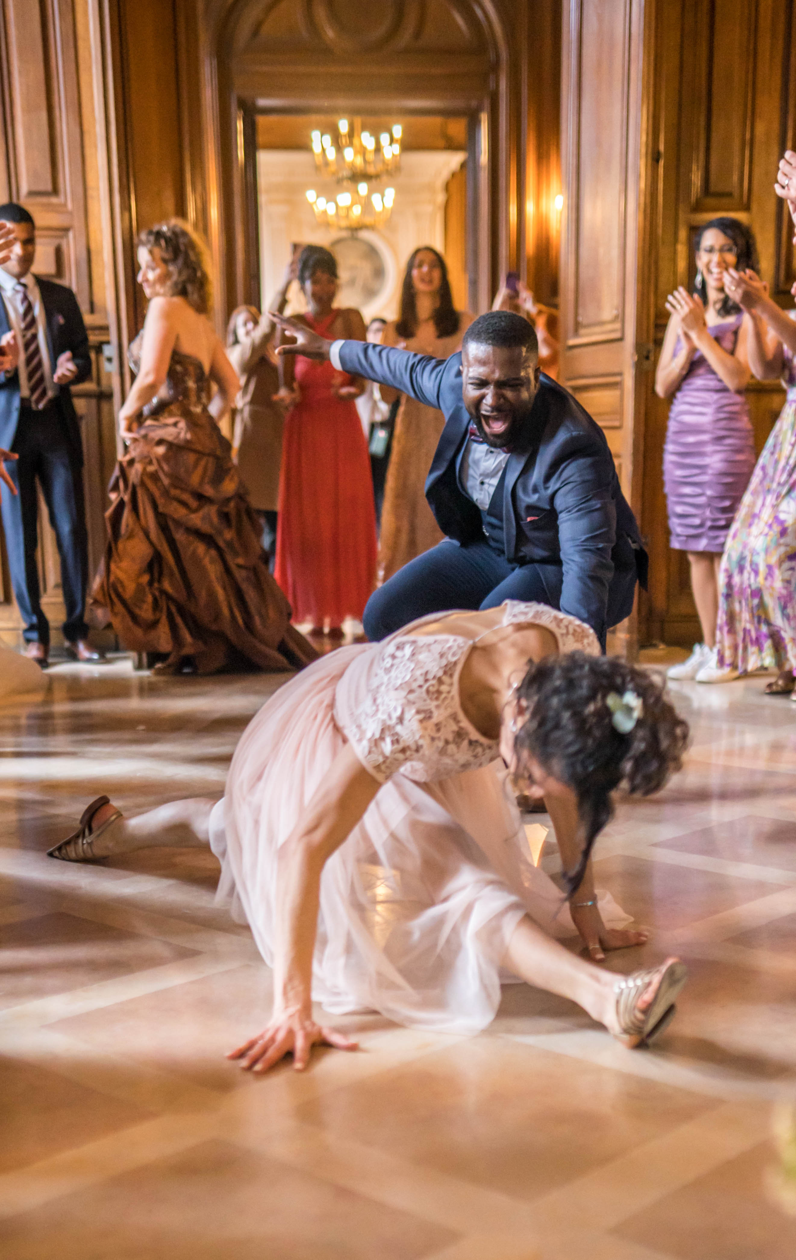 A high-energy reception dance floor moment captured mid-action inside a grand ballroom with warm wood paneling and a chandelier visible in the background corridor. The bride, wearing a blush pink tulle and lace gown with gold sandals and a small floral hair accessory, is low to the parquet floor in a dynamic dance move, while the groom, dressed in a navy blue suit with a light blue shirt, crouches behind her with an expression of joyful excitement. Approximately ten guests line the edges of the dance floor, cheering, clapping, and filming on phones, dressed in a variety of formal attire including a bronze ballgown, a red floor-length gown, a lavender mini dress, and a purple floral dress. The setting appears to be a classic French chateau or hôtel particulier with ornate carved wood boiserie and warm chandelier lighting, and the overall style is classic and formal. Wide shot with a slightly low angle emphasizing the action on the dance floor.