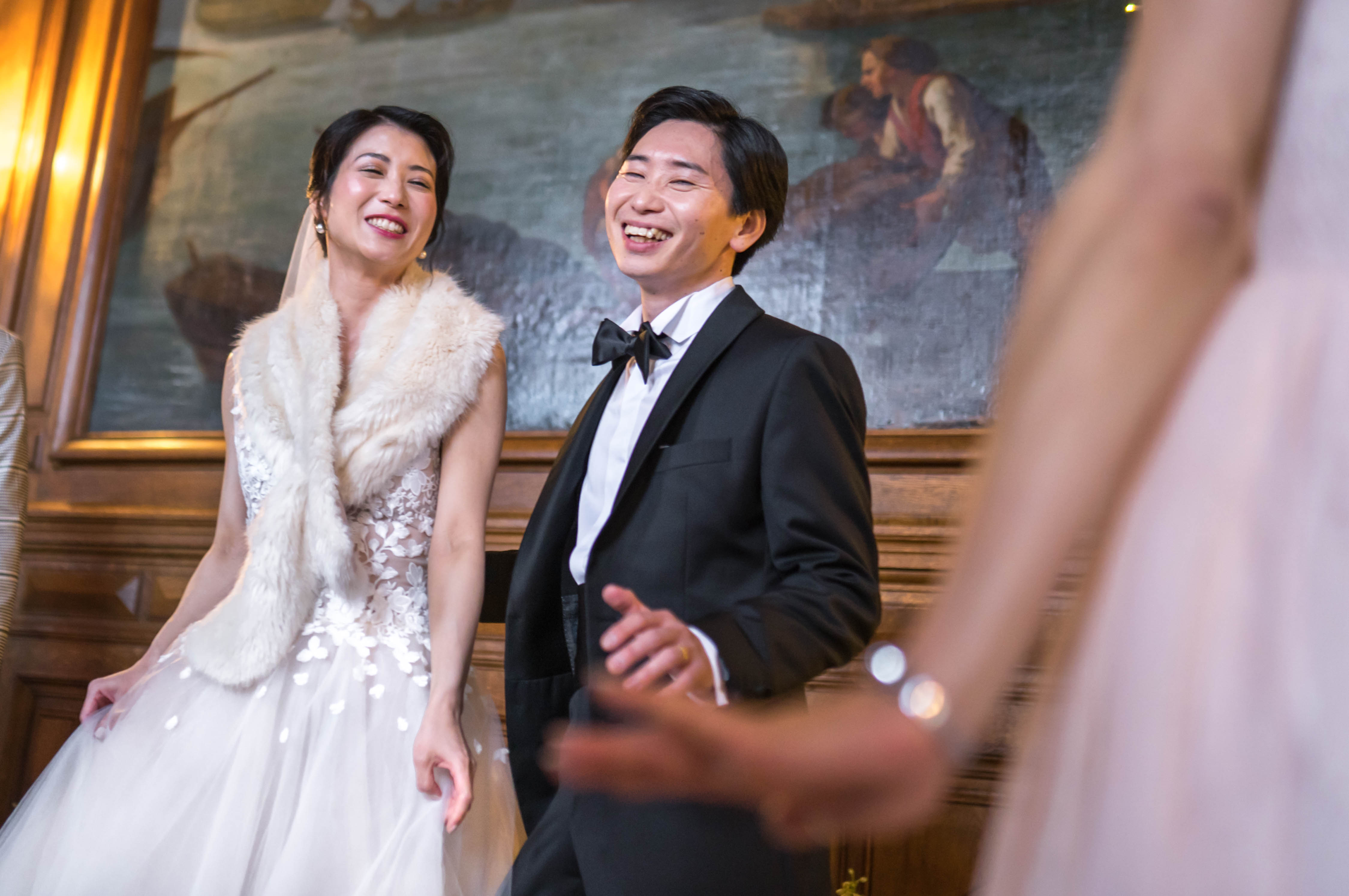 The bride and groom are laughing and reacting to something off-camera, likely during a reception moment such as speeches or a toast, inside a wood-paneled room with a large oil painting on the wall behind them. The bride wears a white floral lace appliqué ballgown with a cream faux-fur stole and a veil, while the groom is dressed in a black tuxedo with a black bow tie. The setting has a classic, formal interior with warm amber wall lighting and dark wood paneling, suggesting a chateau or historic venue. The shot is taken from a low angle with a blurred foreground figure partially visible on the right, giving the image a candid, in-the-moment feel.