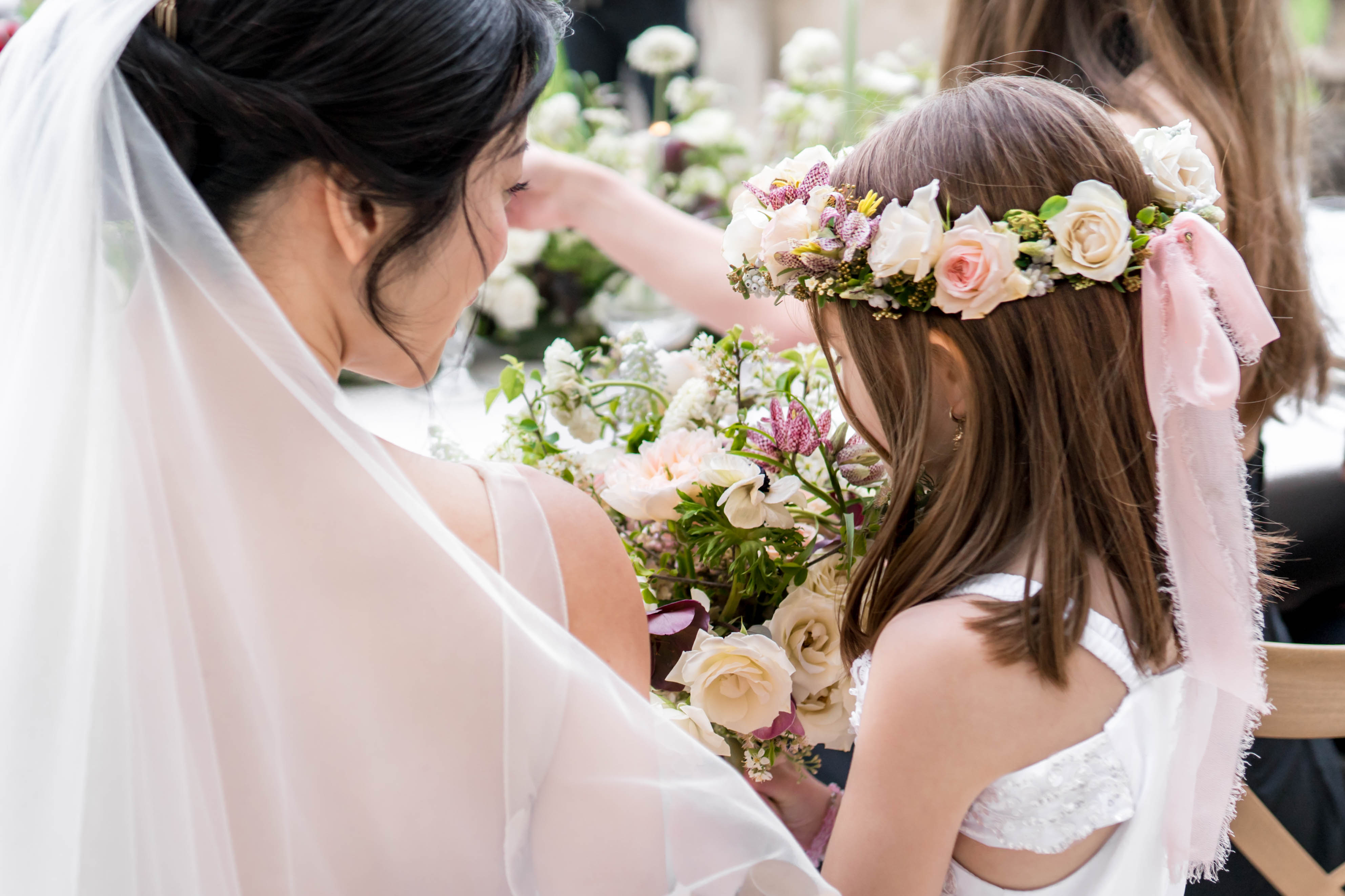A close-up portrait of a bride adjusting the flower crown of a young flower girl before or during the ceremony. The bride has dark hair styled in an updo and wears a white veil, while the flower girl wears a white dress with an open back and a flower crown made of blush pink and ivory roses, small purple astrantia, yellow buds, and greenery, finished with a soft pink ribbon bow. Between them sits a large bridal bouquet featuring cream and blush roses, white astrantia, purple-toned flowers, dark calla lily stems, and lush greenery. Additional floral arrangements are visible in the soft-focus background, consistent with a decorated reception or ceremony space. The overall floral palette is white, blush, and dusty mauve with a garden-style aesthetic.