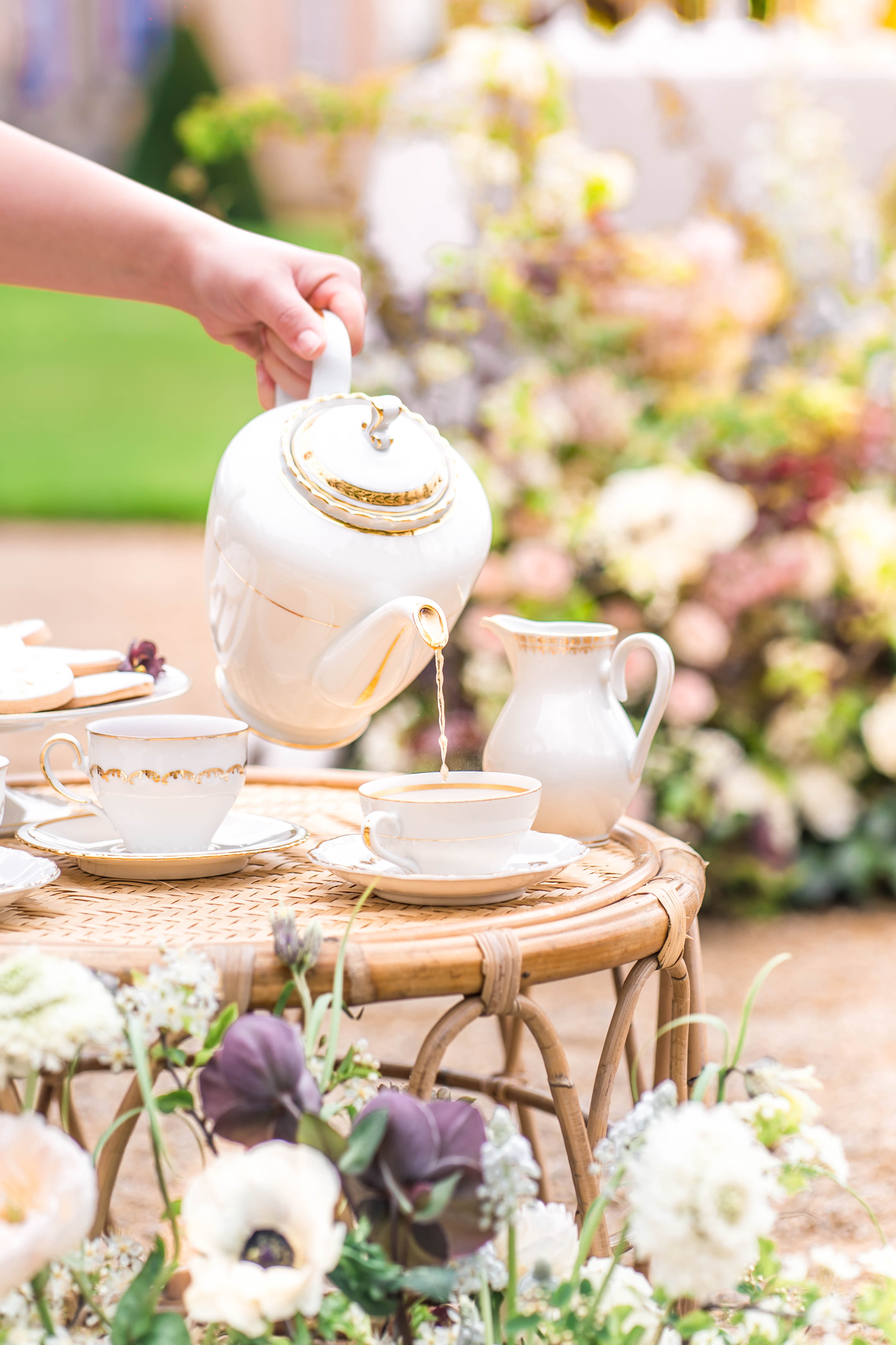 Close-up detail shot of an outdoor wedding tea service, with a hand pouring tea from a white and gold-trimmed porcelain teapot into a matching teacup and saucer set on a round rattan side table. The white porcelain set features gold banding details and includes a small cream pitcher alongside multiple teacups with saucers and a plate of biscuits. The foreground features loose floral arrangements with white anemones, deep purple/aubergine flowers, and greenery, while a large lush floral installation in blush pink, cream, and mauve tones is softly blurred in the background alongside what appears to be a tiered wedding cake. The overall styling is classic with bohemian natural elements, combining vintage-style china with rattan furniture and abundant garden-style florals.
