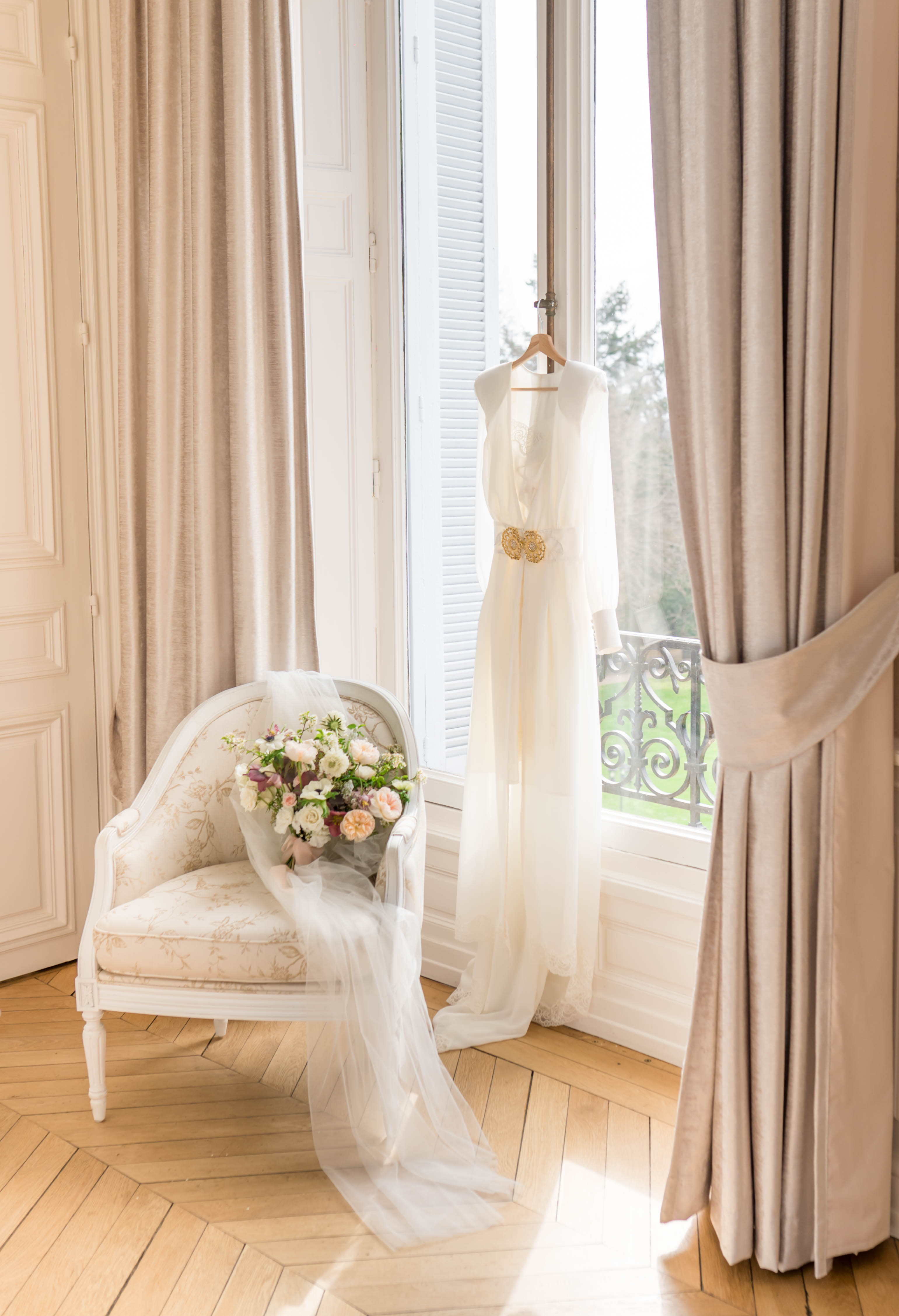 A getting-ready detail shot taken indoors in what appears to be a classic French chateau bridal suite. An ivory sleeveless wedding dress with a gold brooch at the waist and a flowing tulle skirt hangs from a curtain rod in front of tall French doors with white shutters, while a long tulle veil drapes down and pools onto the herringbone parquet floor. A white-painted Louis XVI-style armchair upholstered in a cream floral fabric holds the bridal bouquet, which features blush garden roses, white ranunculus, deep burgundy blooms, and greenery. The room is dressed with floor-length greige linen curtains and the overall decor palette is ivory, cream, and warm taupe with classic French interior styling.