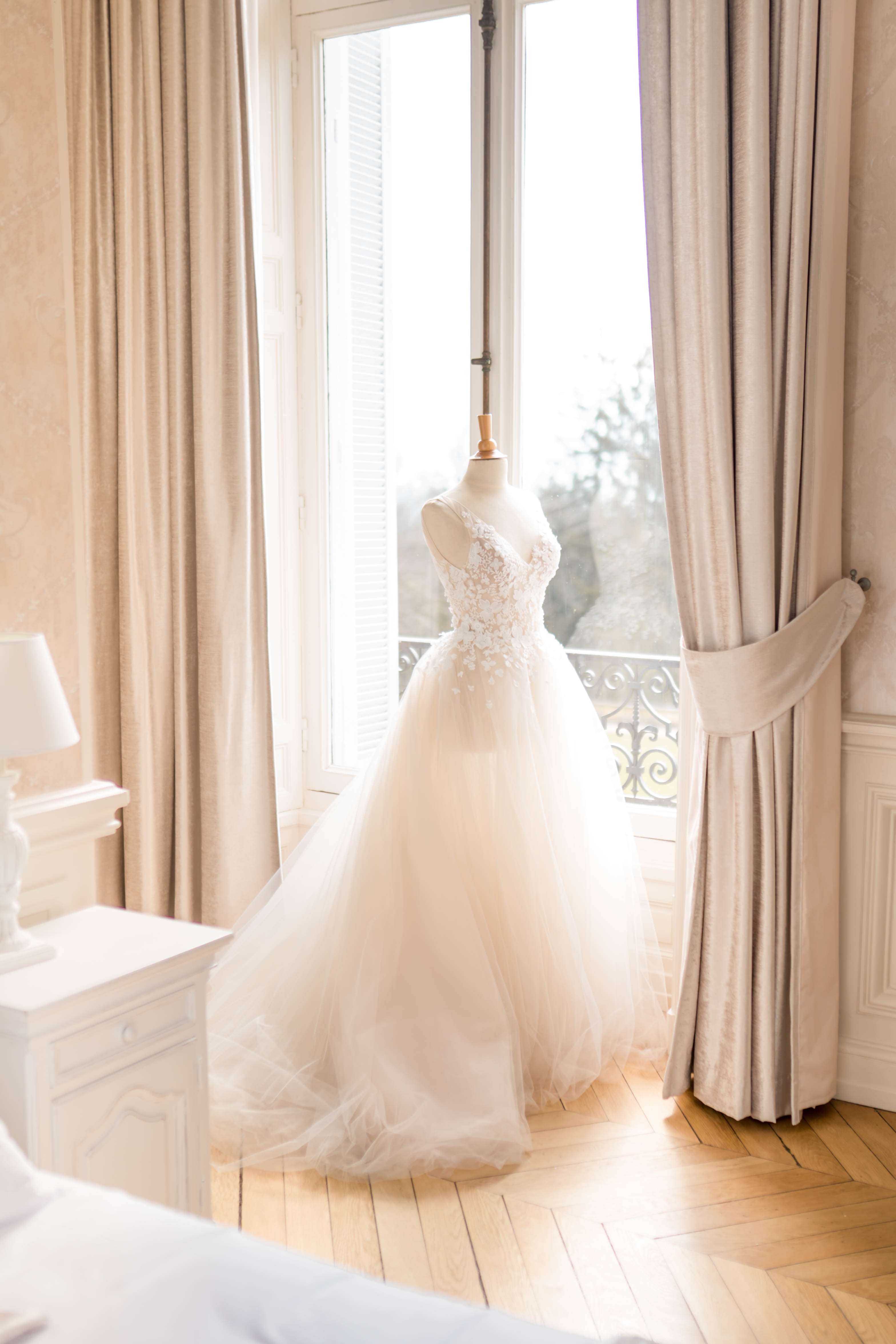 A getting-ready detail shot of a bridal gown displayed on a dressmaker's mannequin, positioned in front of a tall French window with wrought-iron balcony railings visible beyond. The gown features a fitted V-neck bodice with three-dimensional floral lace appliqués in ivory and blush, and a voluminous full-length tulle skirt in champagne-blush tones with a sweeping train spread across the herringbone parquet floor. The room is decorated in a classic French style with floor-length taupe linen curtains, patterned wallpaper, and a white bedside table with a small lamp visible to the left. Natural window light backlit the dress, giving the tulle a soft warm glow against the neutral interior palette.