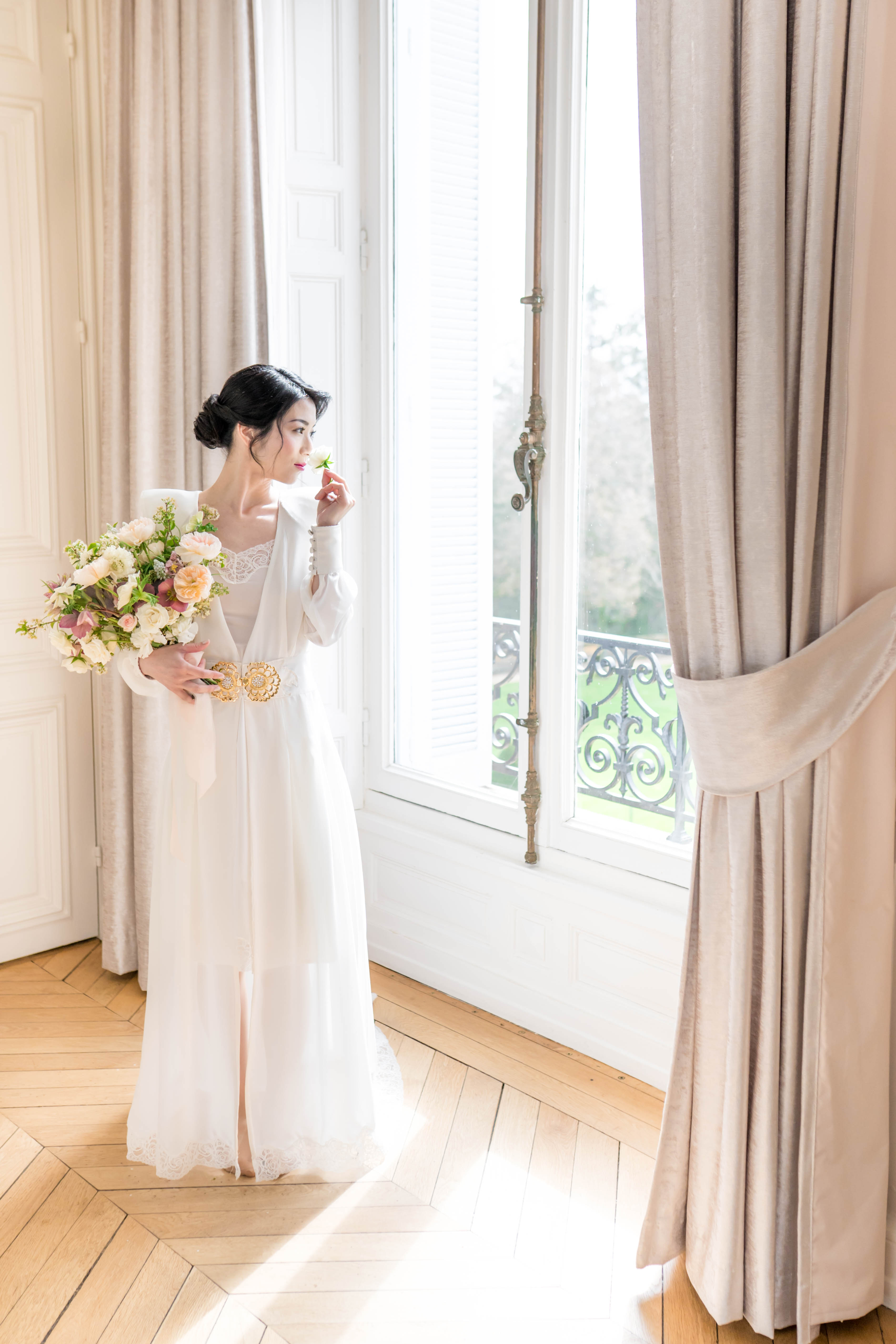A bridal portrait taken indoors in a light-filled room with herringbone parquet flooring, tall white-paneled French doors, and floor-length linen-toned drapes. The bride stands facing the open window, looking to one side, wearing a long-sleeve ivory gown with an off-shoulder neckline, lace hem detail, and a gold medallion belt at the waist. Her dark hair is styled in a low updo, and she holds a loose, garden-style bouquet composed of peach garden roses, blush ranunculus, ivory flowers, and dusty mauve blooms with trailing greenery, while raising a single small white flower toward her face. The composition is a three-quarter-length portrait shot with natural window light illuminating the scene, giving it a classic, airy French interior aesthetic.