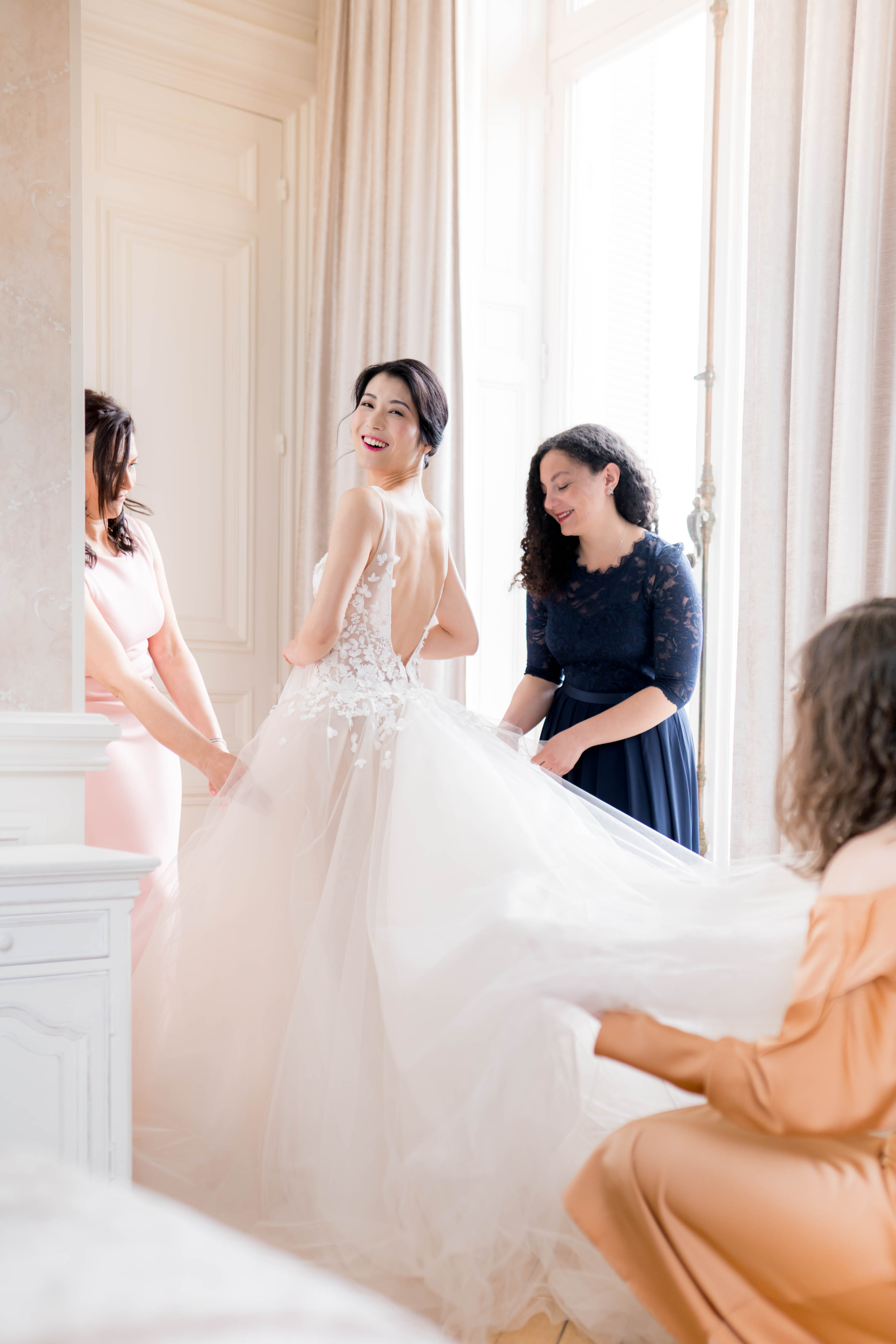 A getting-ready scene captured indoors in a bright, ornate room with white boiserie paneling and floor-length cream curtains, consistent with a French chateau bridal suite. The bride stands at the center, looking back over her shoulder and smiling, wearing a white ball gown with a low open back, 3D floral lace appliqués on the bodice, and a full soft tulle skirt. Three women assist her: one to the left in a blush pink fitted dress holding the train, one behind in a navy blue lace midi dress adjusting the skirt, and one crouching at lower right in a peach/gold outfit spreading the train. The bride's dark hair is styled in a low updo. The composition is a medium portrait shot taken from a slightly low angle, with soft natural backlight from a large window creating a bright, airy atmosphere.