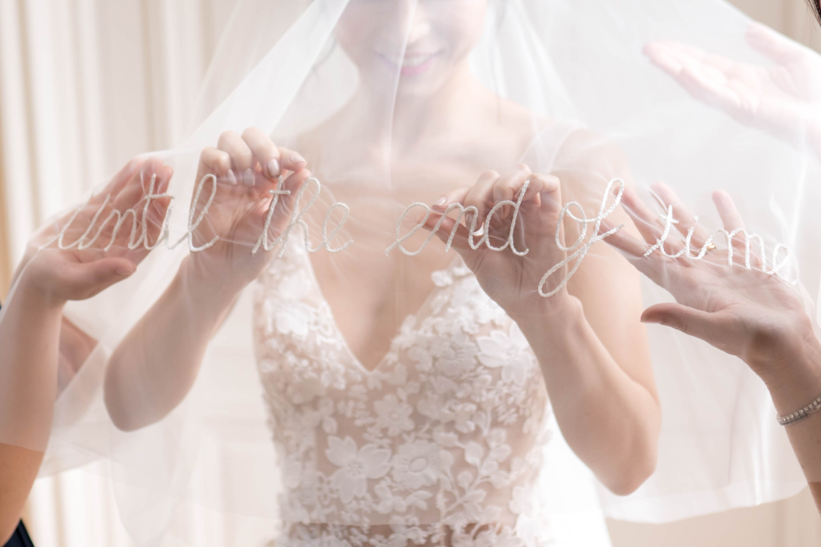 A close-up detail shot taken during the getting-ready portion of the wedding day, showing a bride in a white floral appliqué lace gown with a deep V-neckline holding out her veil to display the phrase 'until the end of time' embroidered in white cursive lettering along its edge. Two additional sets of hands — likely belonging to bridesmaids or helpers — assist in stretching the veil taut so the embroidered text is fully legible. The setting appears to be an indoor preparation room with soft, diffused natural light and neutral cream drapery in the background. The composition is a mid-range portrait cropped at the face, keeping the focus on the veil detail and the lace bodice of the dress.