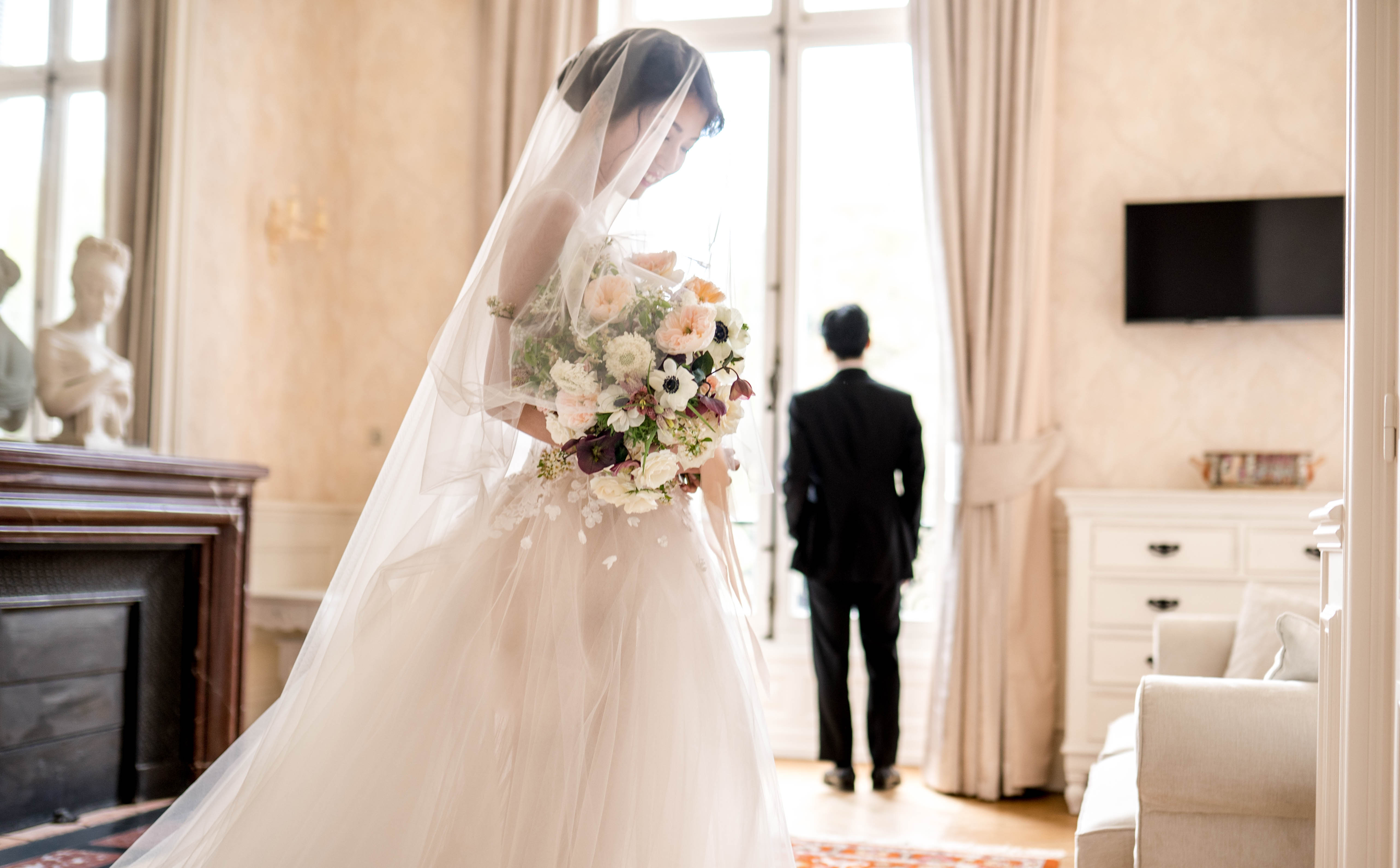 A first-look moment captured indoors in a classically decorated room featuring a fireplace with a marble bust sculpture, tall French doors with floor-length cream drapes, and parquet flooring. The bride stands in the foreground wearing a full-skirted ivory ballgown with floral appliqué detail and a long cathedral-length veil, looking down at her oversized bridal bouquet composed of white anenomes, blush garden roses, ivory scabiosa, deep burgundy calla lilies, and trailing greenery. The groom stands with his back turned toward the French doors, dressed in a black suit, awaiting the reveal. The composition is a medium portrait shot that captures both figures in the same frame while keeping the focus on the bride in the foreground.