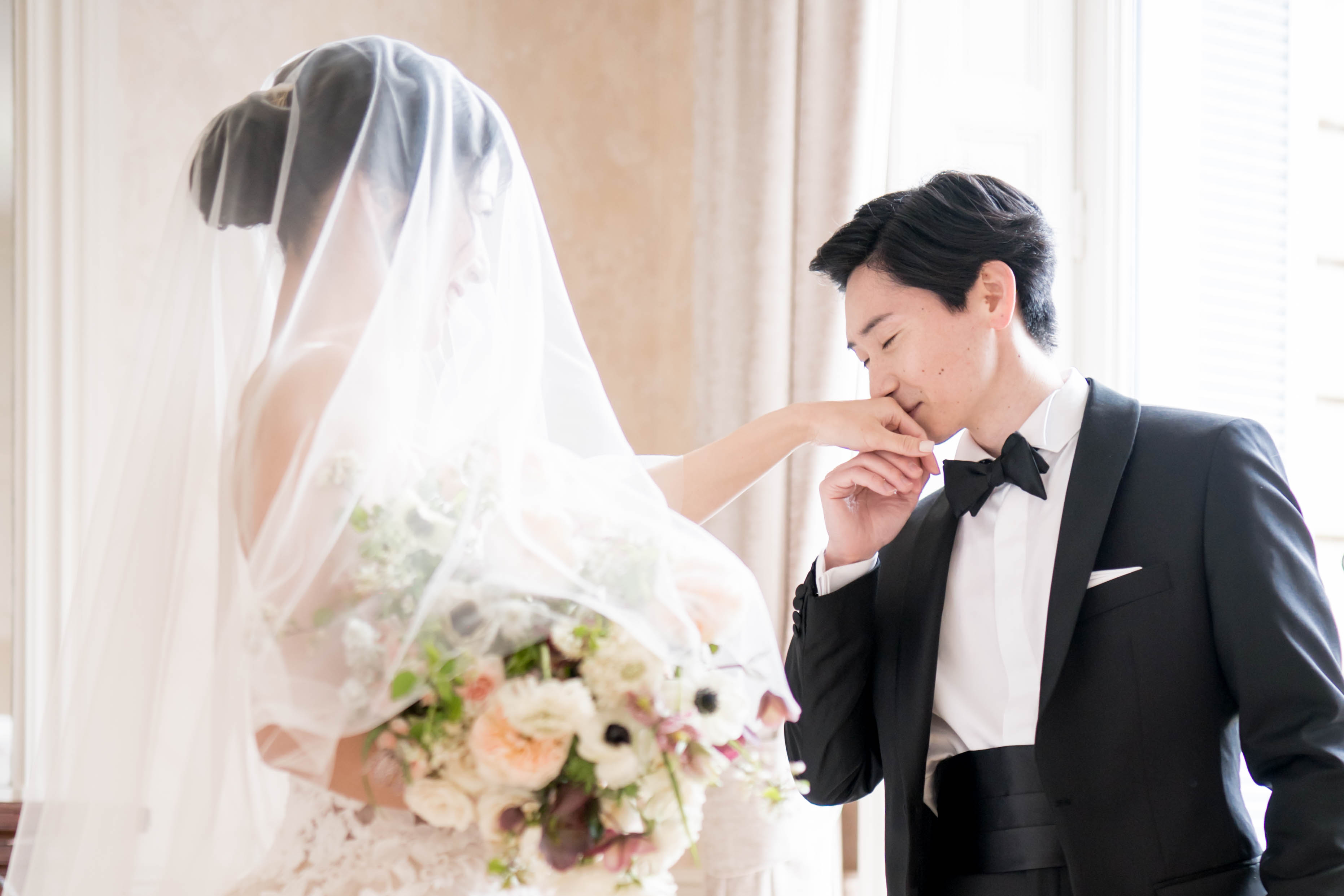 A couple portrait shot indoors, likely during a first look or post-ceremony moment in a bright, ornate interior room with large draped windows. The groom, wearing a black tuxedo with a black bow tie, cummerbund, and white pocket square, is gently kissing the bride's hand. The bride faces away from the camera, wearing a lace wedding gown and a long cathedral-length veil over an upswept hairstyle. She holds a large, lush bouquet featuring blush garden roses, white anemones with dark centers, deep burgundy blooms, and trailing greenery. The composition is a medium portrait shot lit by soft natural window light, with a classic, formal styling aesthetic.