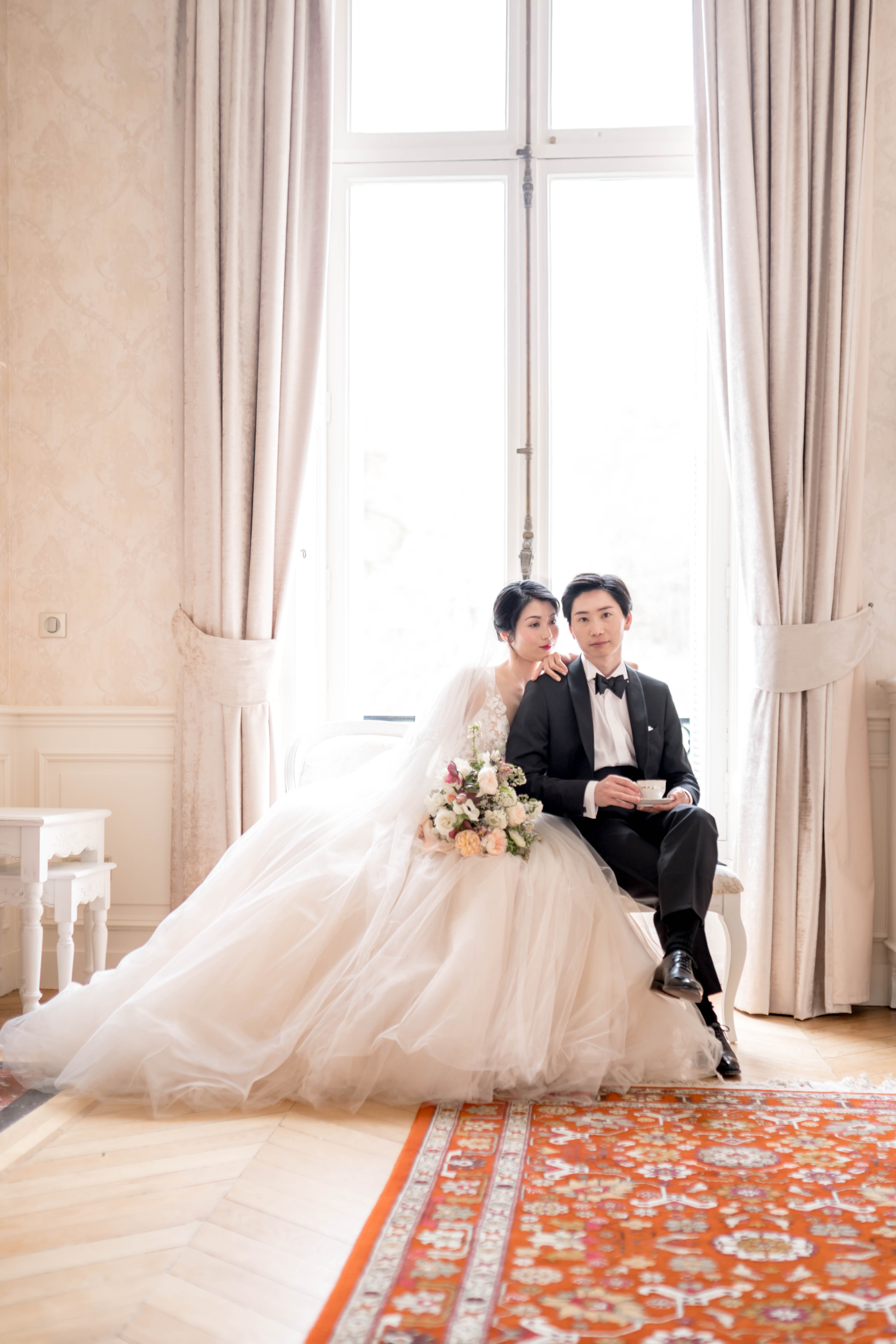 A couple portrait taken indoors in a grand room with tall floor-to-ceiling windows, cream-toned wall paneling with damask detailing, and floor-length taupe curtains. The bride wears a full-volume white tulle ballgown with a lace bodice and holds a loosely arranged bouquet of blush, peach, and dusty pink flowers including what appear to be ranunculus and garden roses with trailing greenery; the groom is dressed in a black tuxedo with a white dress shirt and black bow tie, seated and holding a fine china teacup and saucer. Both look directly at the camera, with the bride standing slightly behind and resting her arm on the groom's shoulder. The room features a parquet wood floor and a bold terracotta-red floral area rug in the foreground, and a small white ornate side table is visible to the left. The overall styling is classic and formal, with a Parisian interior aesthetic. Medium full-length portrait shot with the window providing strong backlight.