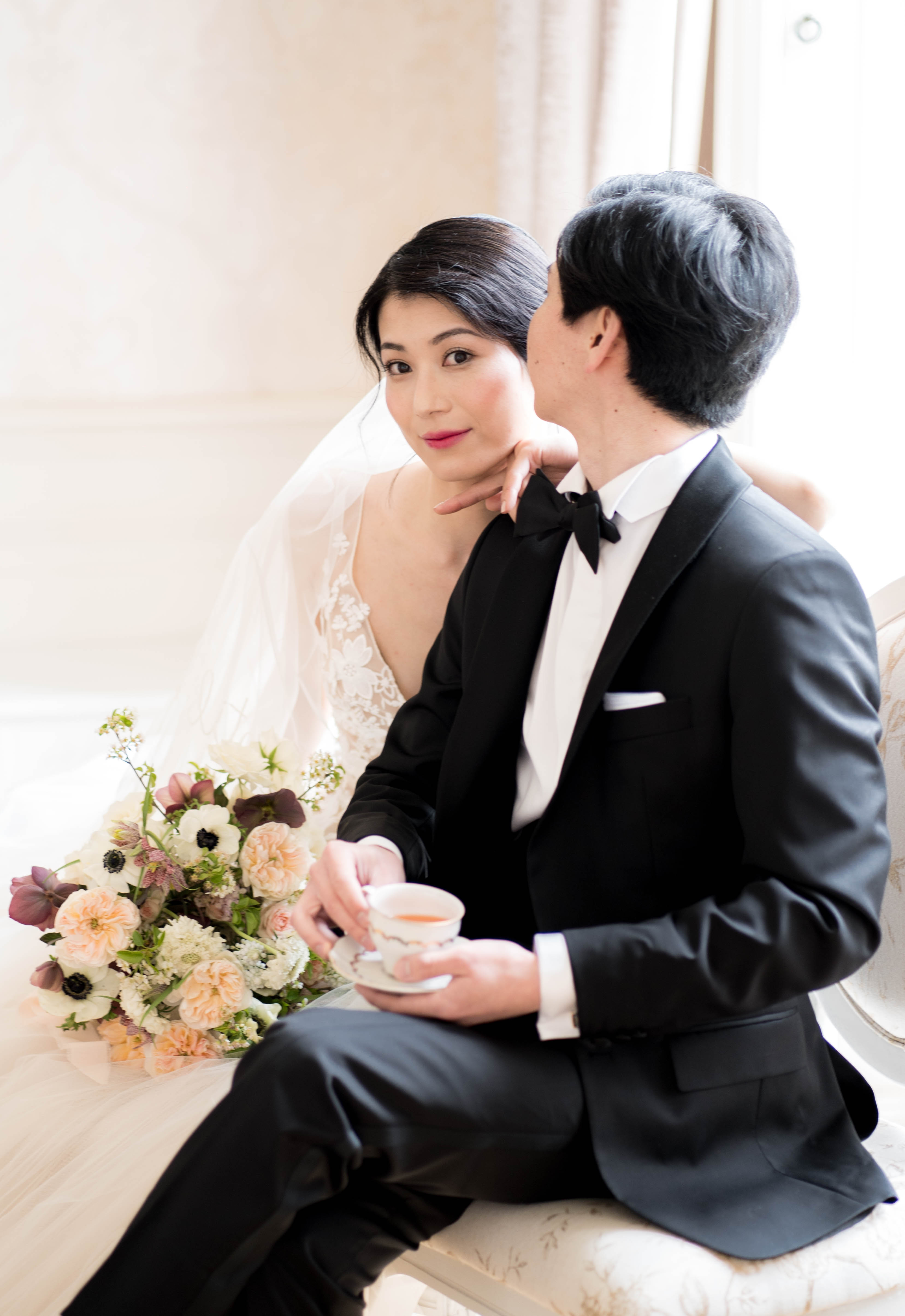 A couple portrait shot indoors, likely in a formal salon or chateau interior with pale cream walls and natural window light. The bride wears a white lace deep-V gown with a sheer veil and has her dark hair pulled back sleekly, while the groom is dressed in a classic black tuxedo with a black bow tie and white pocket square. The groom leans in close to the bride's cheek as she looks directly at the camera, with his hand gently cupping her jaw; he holds a fine china teacup and saucer in his other hand. A loose bridal bouquet rests beside them featuring peach garden roses, white anemones, dark burgundy hellebores, white scabiosa, and delicate trailing stems, creating a soft romantic palette against the dark attire.