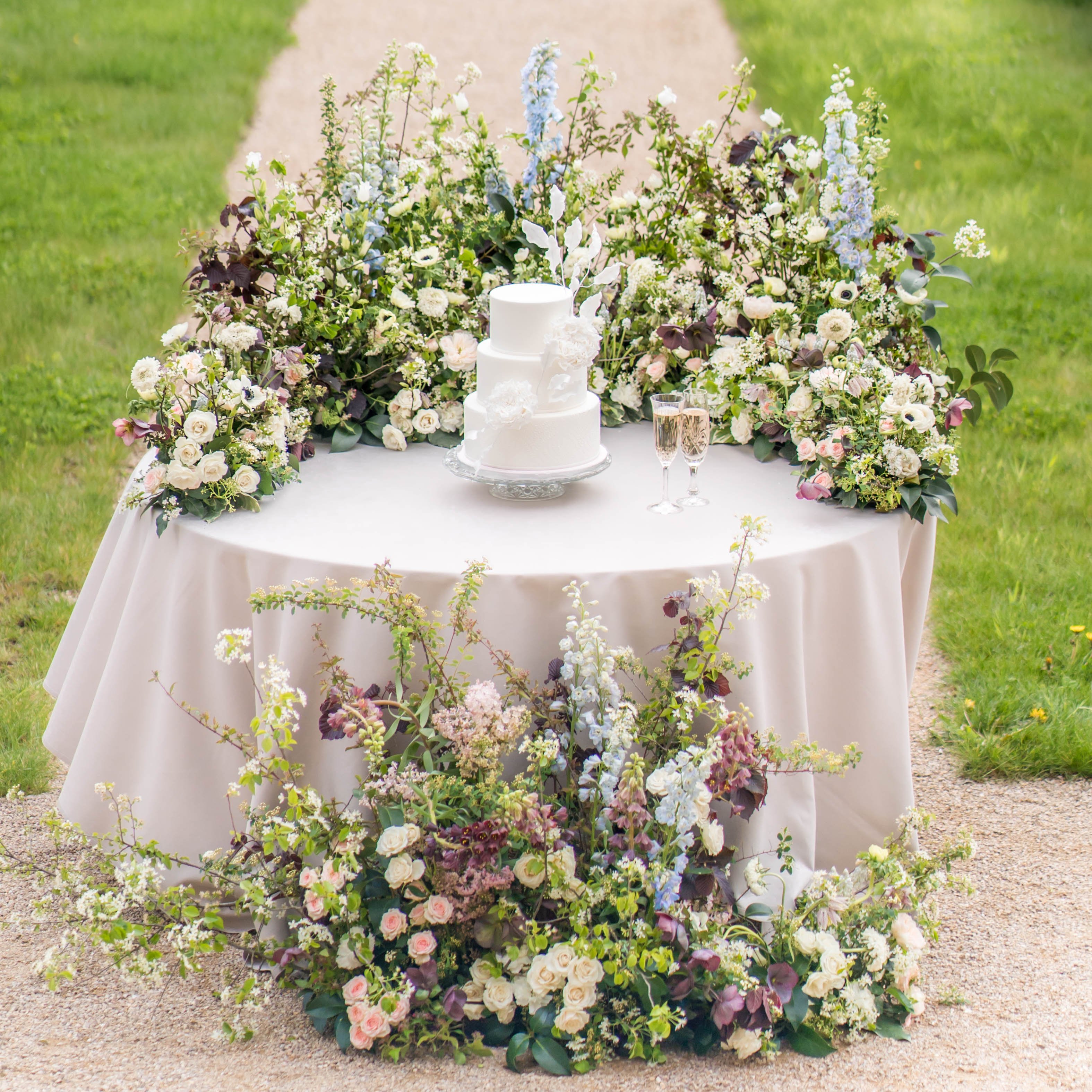 A detail shot of an outdoor wedding cake table set on a gravel path, covered with a blush-ivory satin linen. A three-tier white fondant cake decorated with white appliqué floral details sits on a silver cake stand at the center, flanked by two champagne flutes. The table is surrounded by an abundant, garden-style floral installation that cascades across the tabletop and spills down to the ground, featuring blush garden roses, ivory ranunculus, white anemones, pale blue delphiniums, burgundy hellebores, dusty mauve hydrangea, and trailing green foliage with dark-leafed branches. The floral arrangement wraps entirely around the base of the table as well, creating a full 360-degree effect. The styling palette combines soft blush, ivory, pale blue, and deep plum tones in a romantic garden aesthetic.