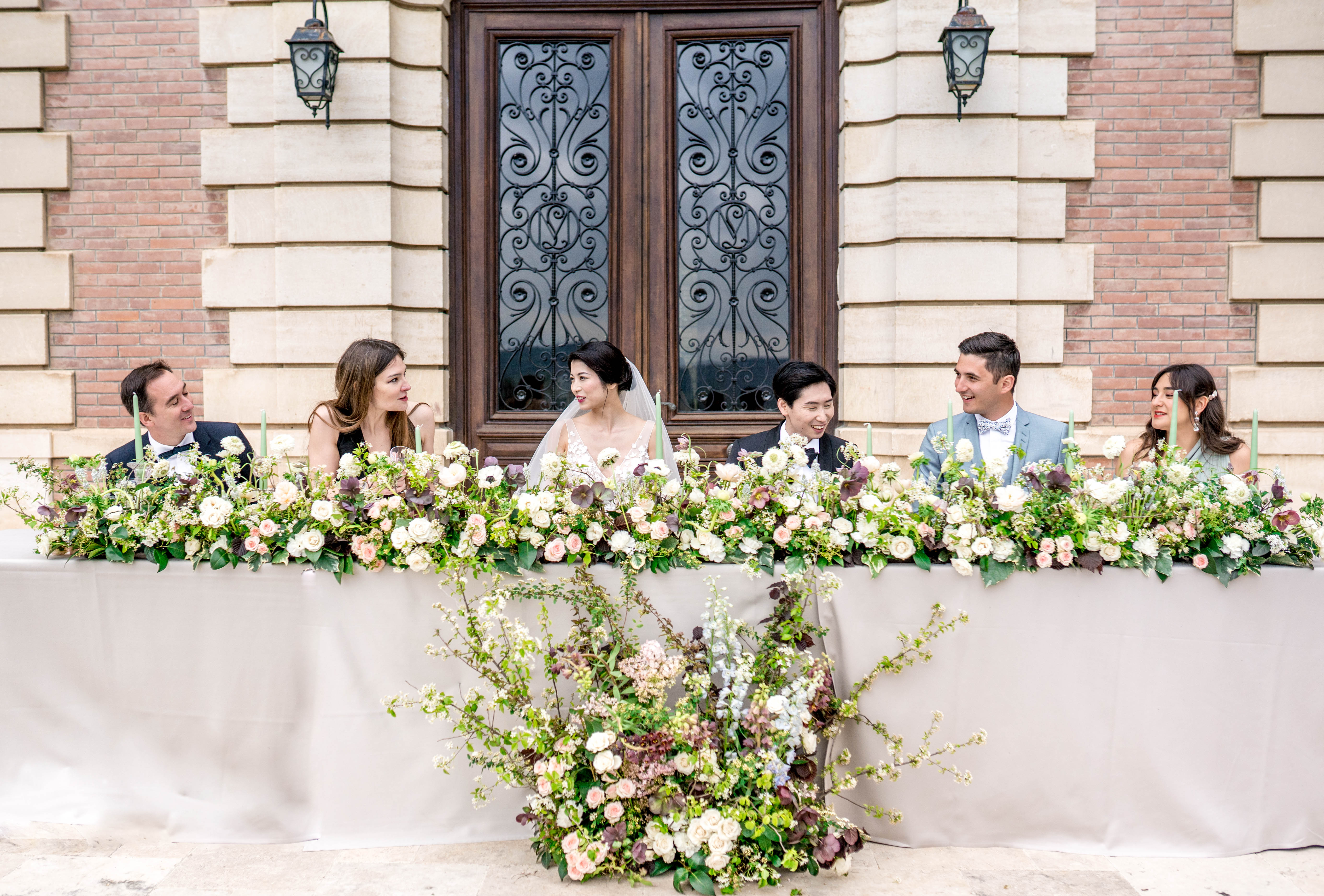 A wedding reception head table scene photographed outdoors against the facade of a chateau-style venue featuring ornate wrought-iron double doors with decorative ironwork monogram detail and wall-mounted lanterns. Six people are seated at a long rectangular table draped in a grey linen: the bride in a white lace gown with a cathedral veil and the groom in a dark suit are centered, flanked by two couples — one man in a navy tuxedo with a woman in a black dress on the left, and a man in a light grey suit with a bow tie and a woman in a sage green dress on the right. The table is lined with a dense, lush floral garland featuring ivory garden roses, blush roses, white ranunculus, dark burgundy/chocolate cosmos, and abundant greenery, with thin sage green taper candles interspersed throughout. A large cascading floral arrangement spills down the front-center of the table onto the ground, incorporating flowering branches, blush and ivory blooms, and trailing greenery. The overall decor palette is blush, ivory, burgundy, and green with a classic, garden-inspired styling. Wide shot taken straight-on at table height.