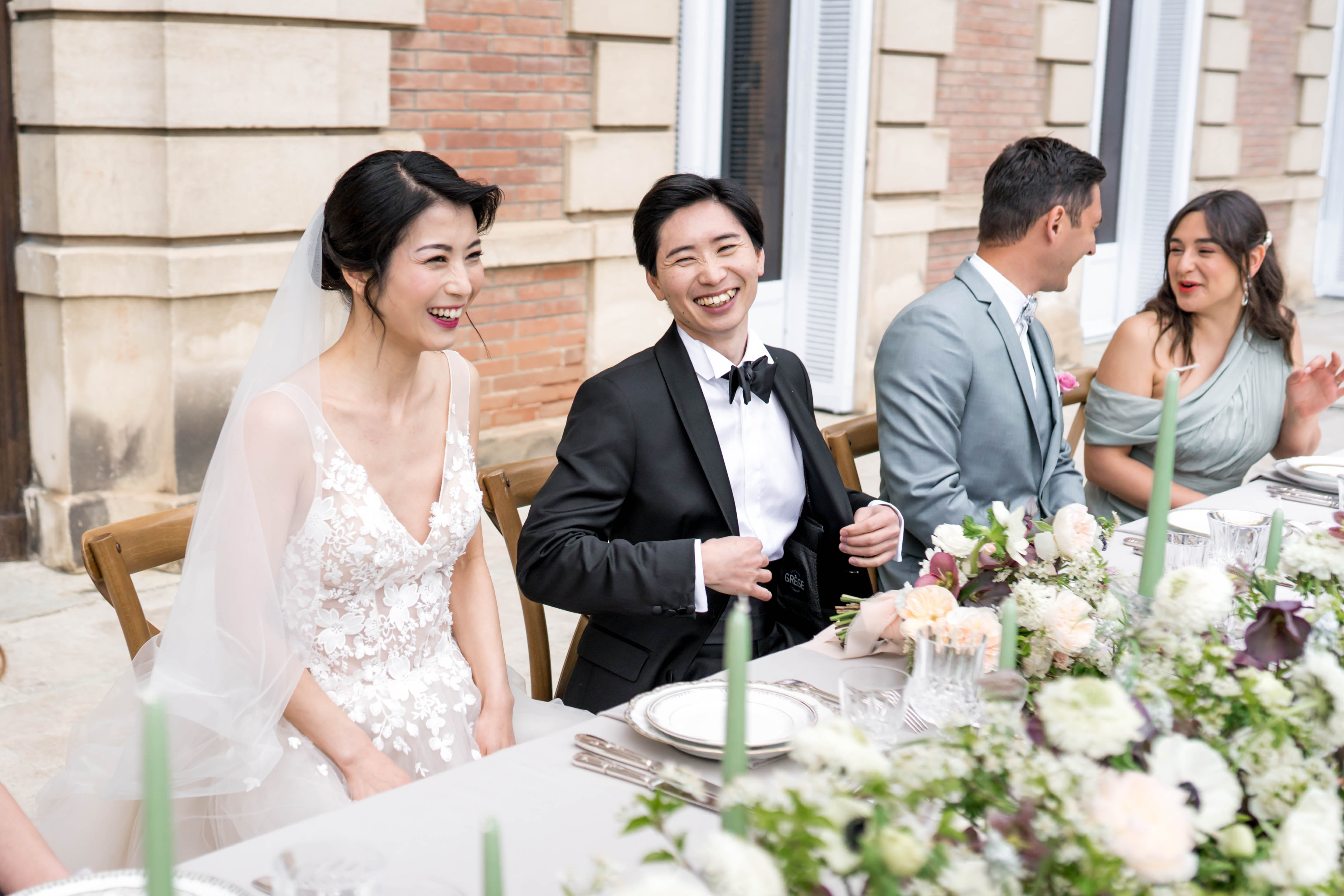 The bride and groom are seated at a reception dining table outdoors, both laughing, likely during a speech or toast. The setting appears to be a terrace or courtyard adjacent to a building with brick and stone facade architecture. The bride wears a white lace appliqué V-neck gown with a sheer veil, and the groom is dressed in a black tuxedo with a black bow tie; two additional guests are visible in the background — a man in a grey suit and a woman in a sage green off-the-shoulder dress. The table is styled with a grey linen, silver-rimmed charger plates, tall sage green taper candles, and a lush floral arrangement featuring ivory ranunculus, blush garden roses, white anemones, and deep plum/mauve blooms with green foliage. The overall decor palette is soft and neutral with sage green accents. Medium shot capturing the couple and nearby guests from a slightly elevated angle.