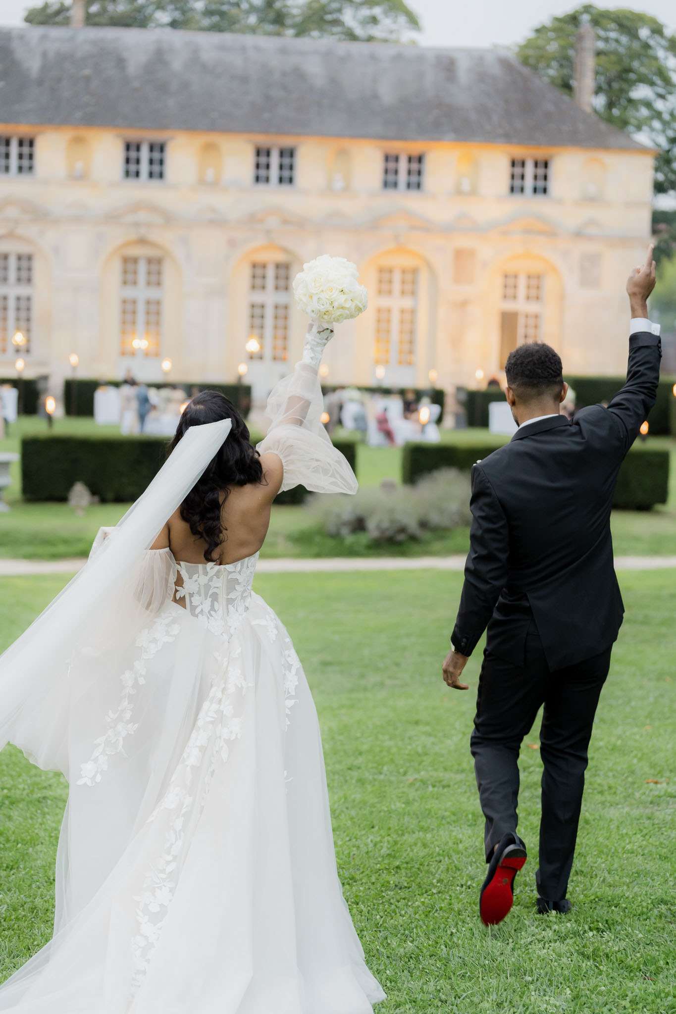 The bride and groom are photographed from behind, walking across a lawn toward a grand French chateau at dusk, both with their arms raised in celebration. The bride wears a white off-shoulder ball gown with floral lace appliqué detailing and sheer puff sleeves, paired with a long white veil and long dark wavy hair; she holds a tightly packed round bouquet of white roses or hydrangeas aloft in one hand. The groom wears a black suit with black trousers and notably red-soled shoes, his right arm raised in the air. In the background, guests mingle on the chateau's formal garden terrace, which is lit with warm candlelight and lanterns against the golden limestone facade of a classic French chateau, with neatly trimmed hedgerows framing the grounds. This is a wide portrait shot capturing a joyful, celebratory moment with a modern classic styling. Potential venue feature image.