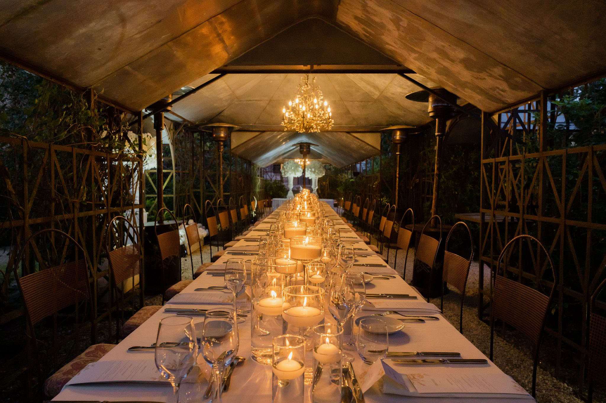 A wide-angle shot of a wedding reception dinner setup inside a covered outdoor structure with an ornate wrought-iron frame and canvas roof, photographed at night before guests are seated. A single long banquet table runs the full length of the structure, dressed in a white linen tablecloth and set with crystal wine glasses, silver flatware, white folded napkins, and printed menus at each place setting. The centerpiece consists of a continuous row of pillar candles in glass cylinders and votive candles, casting warm golden light along the entire table. Wicker and iron bistro-style chairs line both sides of the table, accommodating approximately 30–40 guests. A crystal chandelier hangs at the center of the structure, and patio heaters are positioned along the sides. At the far end, a large white floral arrangement — likely white hydrangeas or similar blooms — serves as a focal backdrop. The overall decor palette is warm ivory and gold with a classic, candlelit garden-party aesthetic. Potential venue feature image.