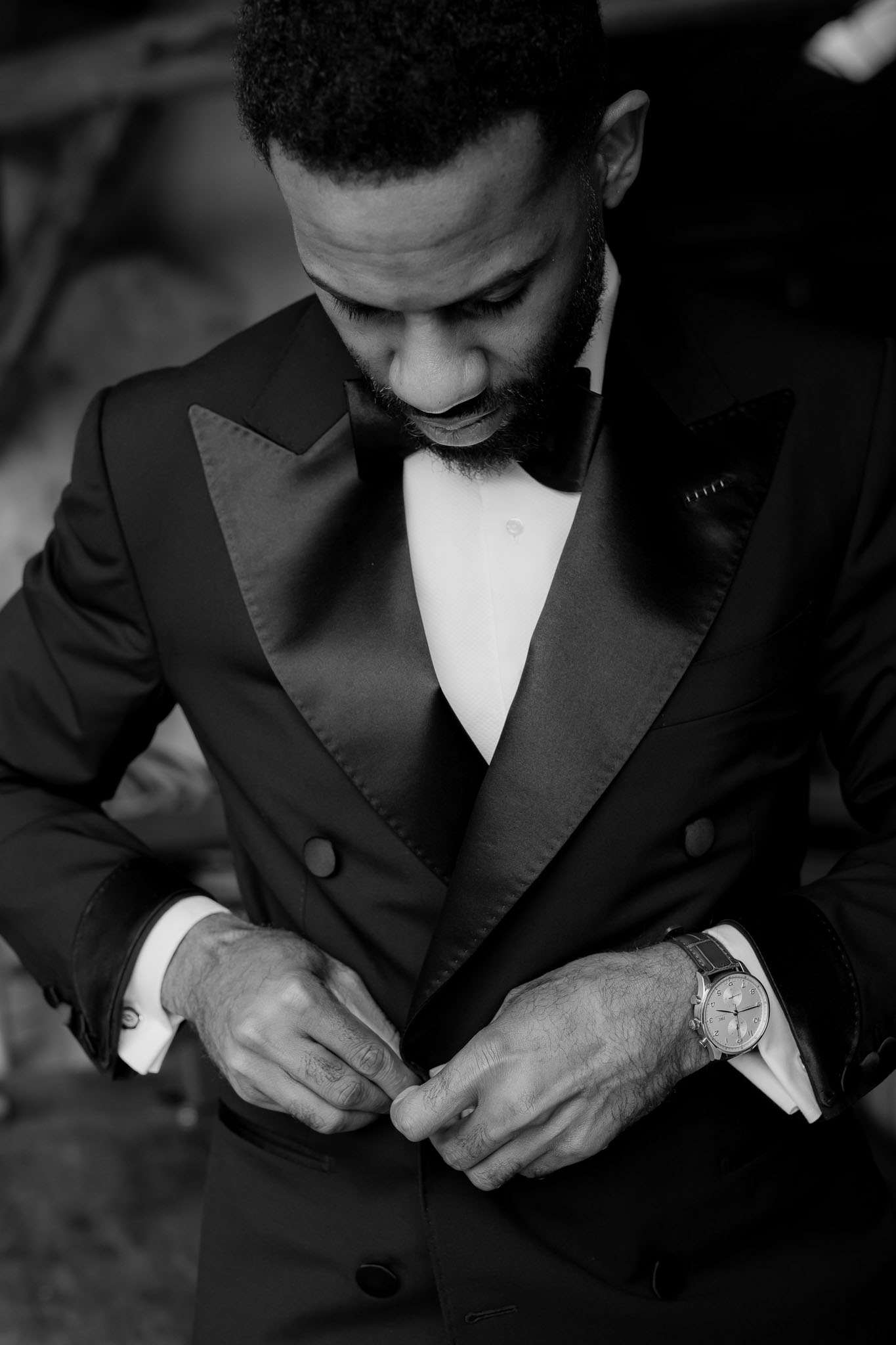A black-and-white close-up portrait of a groom getting ready, captured from a slightly elevated angle as he buttons his double-breasted tuxedo jacket. He is wearing a black tuxedo with satin peak lapels, a white dress shirt with French cuffs, a black bow tie, and a dark pocket square; a dress watch with a leather strap is visible on his left wrist. The image features strong contrast between the white shirt and dark jacket, with the background softly out of focus. The composition is a tight portrait emphasizing the groom's hands and upper body during a candid getting-ready moment.