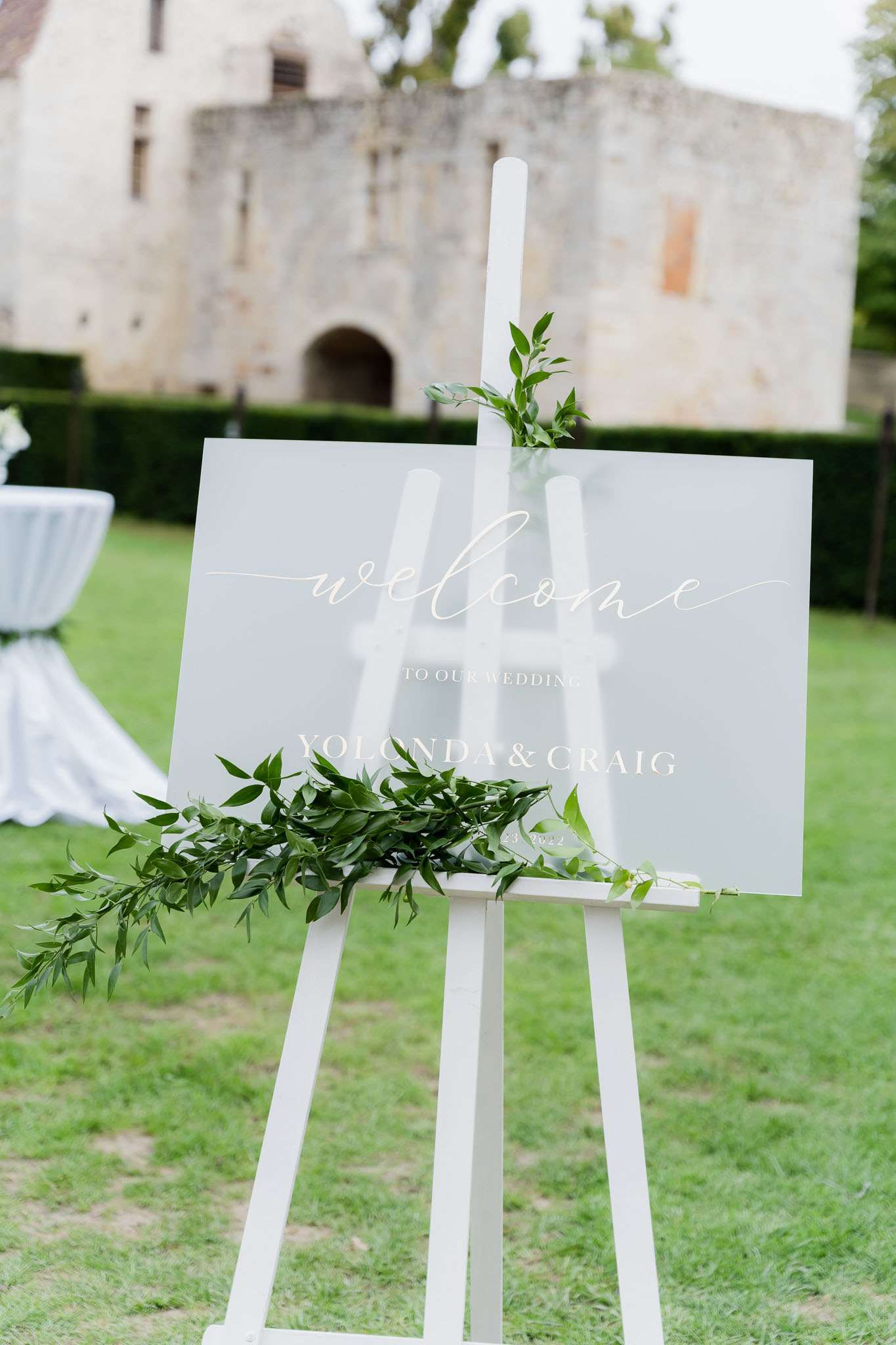 A close-up detail shot of a frosted acrylic wedding welcome sign reading 'Welcome to our Wedding – Yolonda & Craig' in white calligraphy script, displayed on a white wooden easel outdoors on a lawn. The sign is decorated with trailing green foliage — ruscus or similar leafy greenery — draped along the bottom and top of the easel. In the background, a medieval stone chateau tower and a white-draped cocktail table are softly out of focus. The overall decor palette is white and green with a clean, classic styling approach. Potential venue feature image.