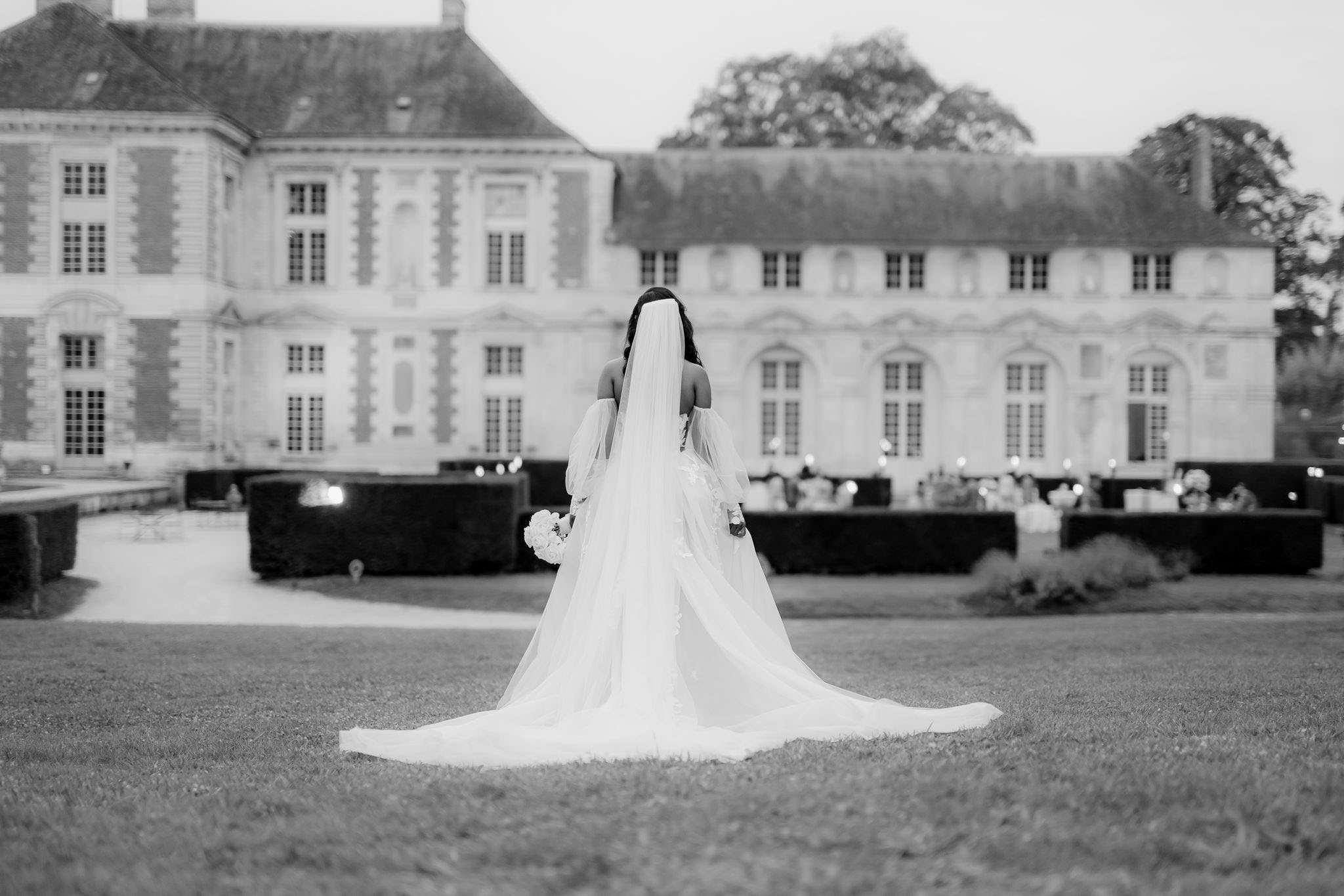 This black-and-white bridal portrait shows a bride standing alone on the lawn of a large French château, photographed from behind. She wears an off-shoulder gown with detached long sleeves, a full cathedral-length train, and a long veil that falls to the ground, both spreading wide across the grass. She holds a round bouquet of what appear to be light-toned flowers, likely peonies or roses. In the background, a formally arranged garden with trimmed hedges is set up for a reception, with candlelight and floral centerpieces visible on tables, and globe lighting accents along the hedgerows. The image is a wide portrait shot with soft mid-tones and gentle contrast, emphasizing the sweep of the gown and veil against the grand classical château façade. Potential venue feature image.