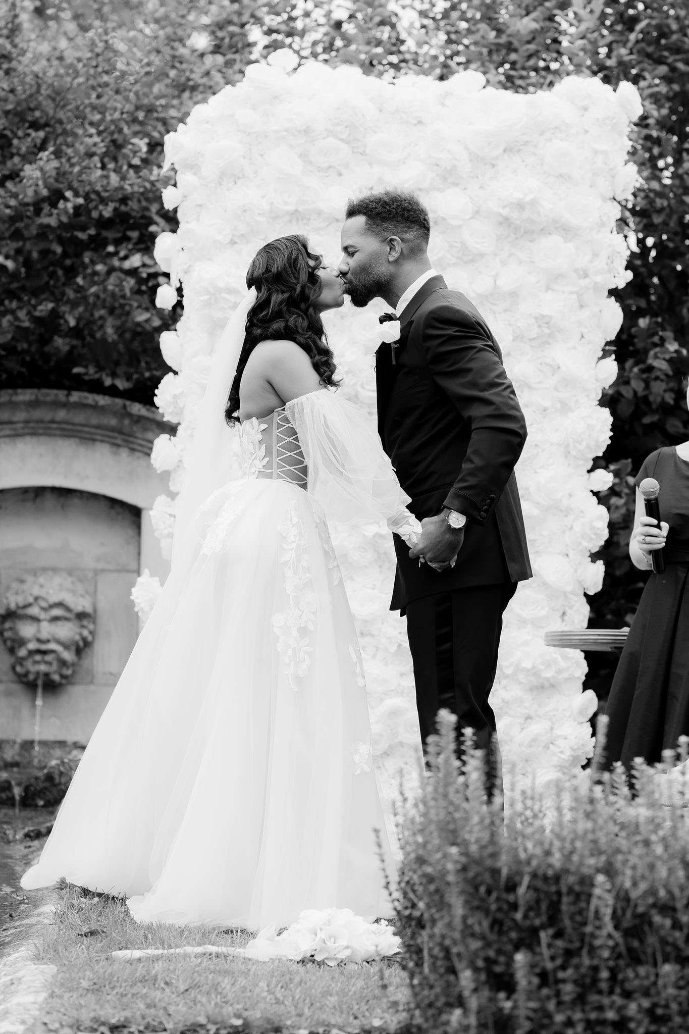 Black-and-white image capturing the first kiss moment during an outdoor wedding ceremony. The bride wears a ball gown with a corset bodice featuring lace-up back detailing, floral appliqué, sheer cape sleeves, and a full tulle skirt; her long dark hair falls in loose waves. The groom wears a dark tuxedo with a bow tie and boutonnière. They are framed by a large rectangular floral wall backdrop densely covered in roses, rendered in bright whites against the dark foliage behind. A stone wall feature with a carved face fountain is visible to the left, suggesting a formal garden or chateau grounds setting. An officiant holding a microphone is partially visible to the right edge of the frame. The composition is a mid-range portrait shot taken from slightly behind and to the side of the couple.