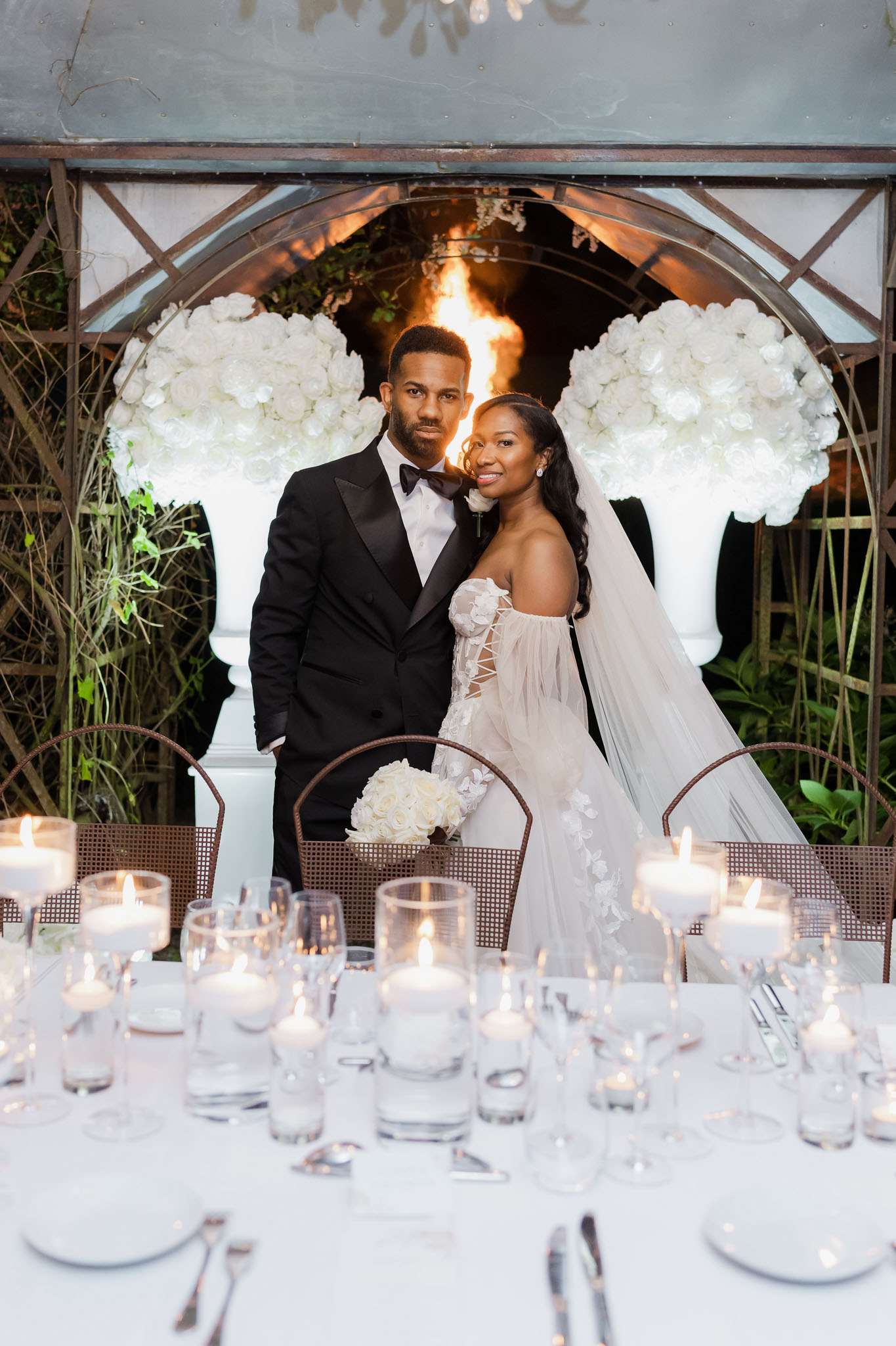 A couple poses together at their wedding reception in an outdoor garden setting, standing behind a white linen-covered sweetheart table set with numerous lit pillar candles in glass vessels, crystal stemware, and silverware. The groom wears a black tuxedo with satin lapels and a black bow tie, while the bride wears a strapless ivory gown with floral appliqué detailing, a corset-style bodice, off-shoulder tulle sleeves, and a long cathedral veil; she holds a round bouquet of white hydrangeas and roses. Behind the couple, a wrought-iron arched structure frames two large white pedestal urns overflowing with white roses and hydrangeas, with an open flame visible between them creating a warm amber glow. The overall decor palette is white and black with warm candlelight, reflecting a modern classic styling theme, captured in a mid-range portrait composition.